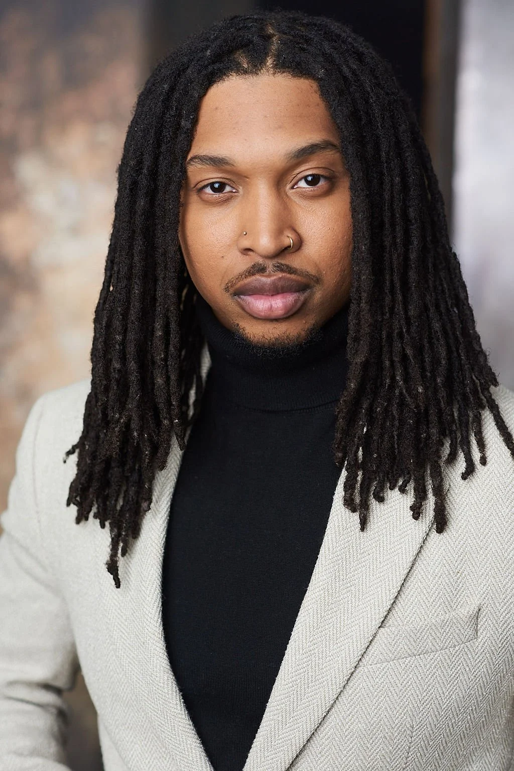 A portrait of a Black man with long dreadlocks, wearing a black turtleneck and a light-colored blazer, standing indoors with a blurred background.