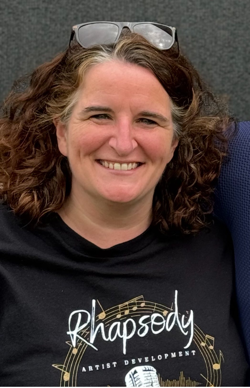 A woman with curly brown hair and sunglasses on her head, smiling in front of a black background, wearing a black T-shirt with a music-themed logo that reads 'Rhapsody Artist Development'.