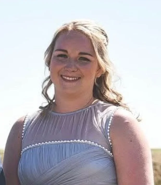 A woman outdoors wearing a sleeveless light purple dress, smiling with blonde hair and a bright background.