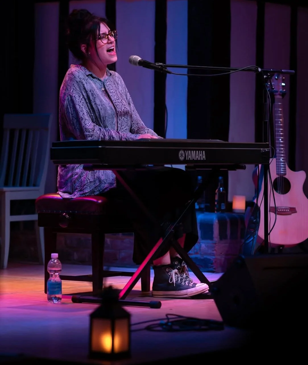 Woman singing and playing keyboard on stage with guitar in background, microphone, and candlelit lantern in foreground.