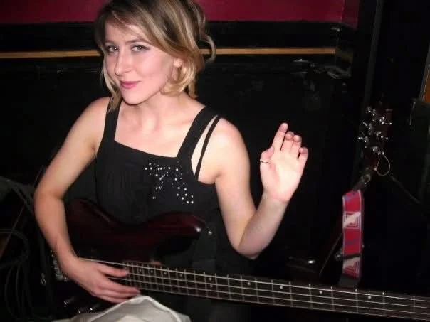 Young woman waving and holding a guitar, in a music studio with audio equipment in the background.