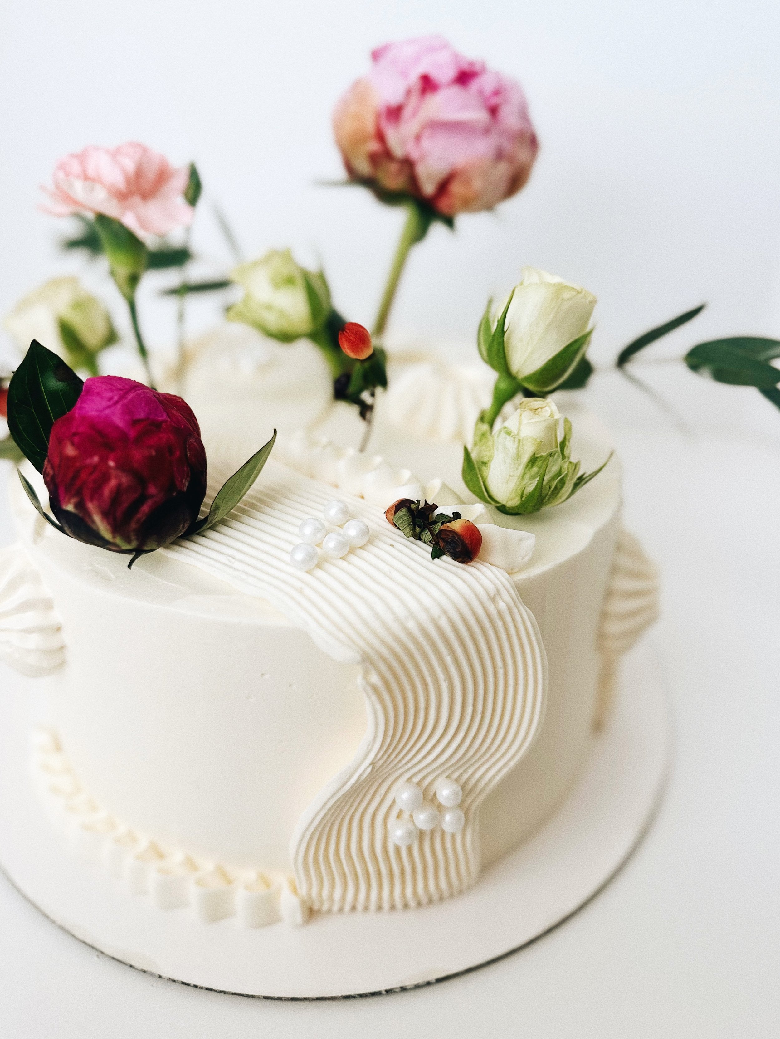 A white cake decorated with pink, red, and white flowers, green leaves, white pearls, and intricate cream piping details.
