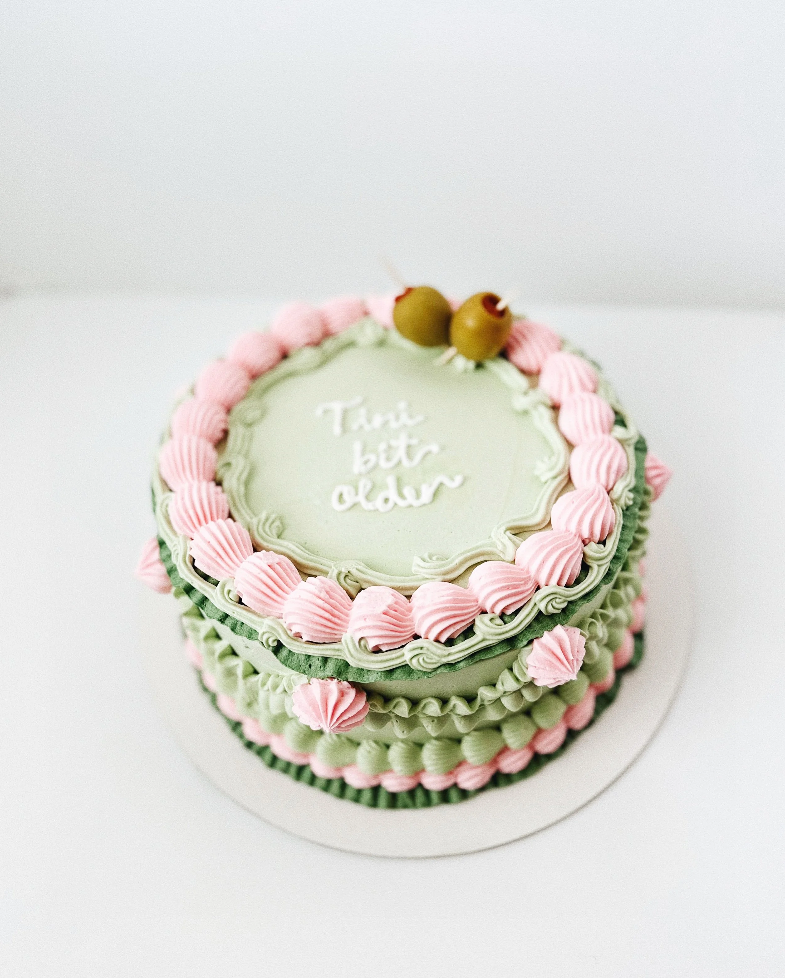 A decorated birthday cake with pink and green frosting and two green olives on top. The cake has the message "Tārei biti older" written on it.