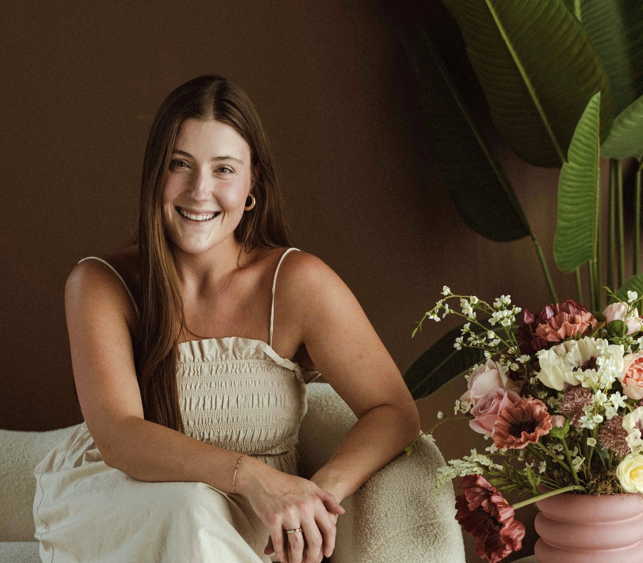 A smiling woman with long brown hair sitting on a cream-colored sofa next to a pink vase with a colorful bouquet of flowers, including pink, white, and red blooms, and green foliage, in a room with brown walls.
