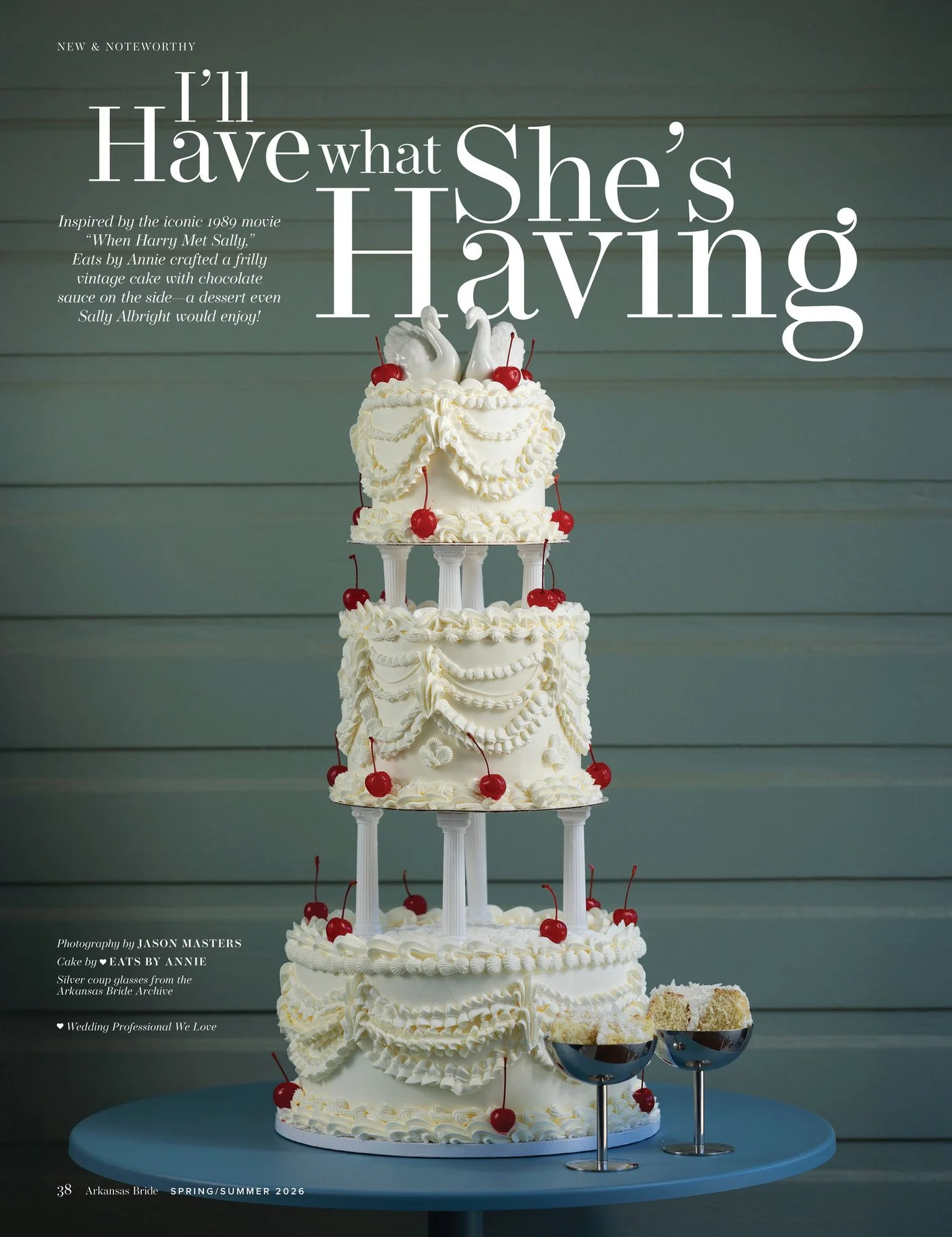 A tall, layered wedding cake with white icing decorated with cherries and swan toppers, displayed on a blue table, with cake slices in silver cups.