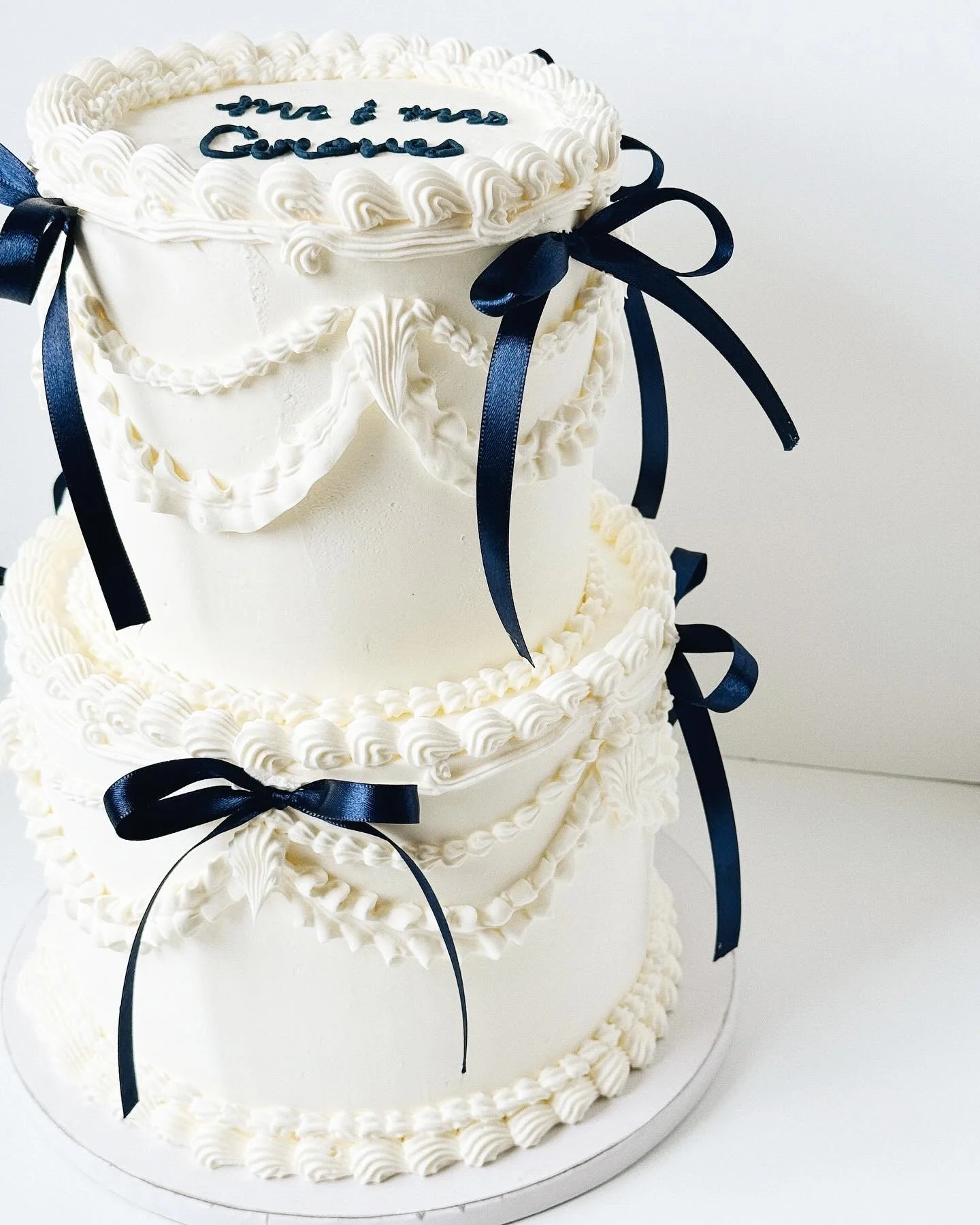 Three-tier white wedding cake decorated with white icing rosettes and bows tied with navy blue ribbons. The top tier has a message written in navy blue icing saying 'you & me forever'.