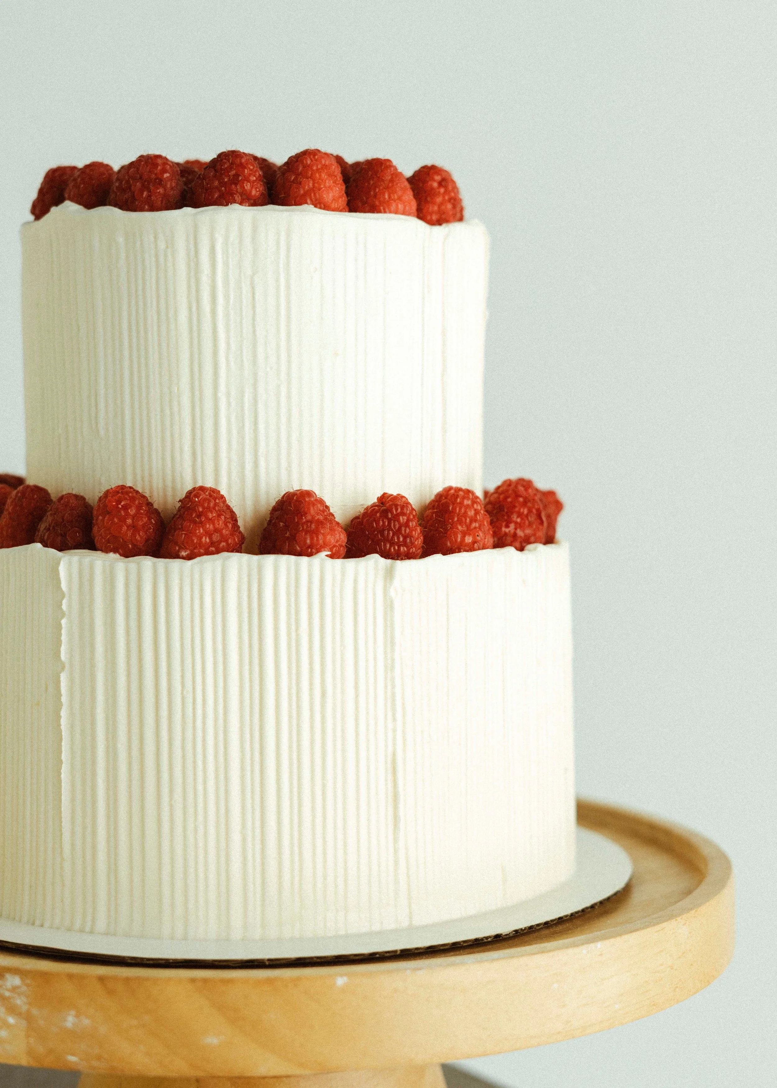 A two-tiered white birthday cake decorated with raspberries on the top and middle edges, placed on a wooden cake stand.