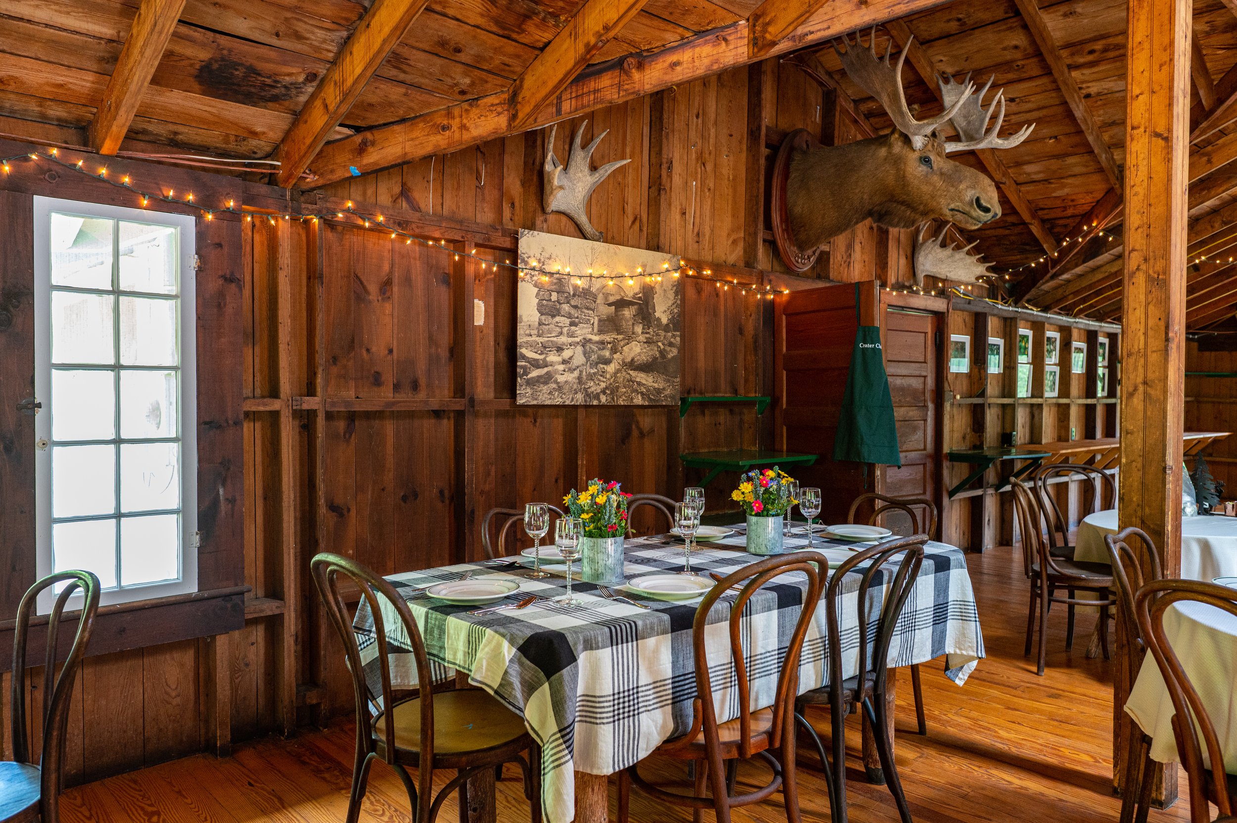 A rustic restaurant with wooden walls and ceiling, decorated with mounted deer heads and string lights. A table is set with a checkered tablecloth, plates, glasses, and a flower centerpiece.