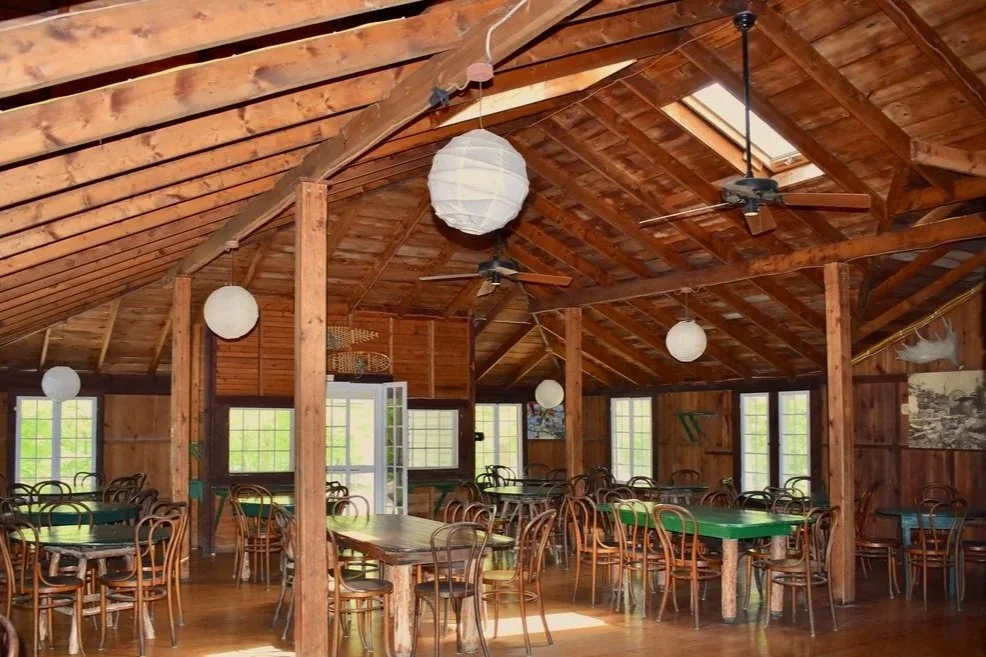 A rustic wooden dining area with several tables and chairs, hanging paper lantern lights, ceiling fans, and multiple windows letting in natural light.
