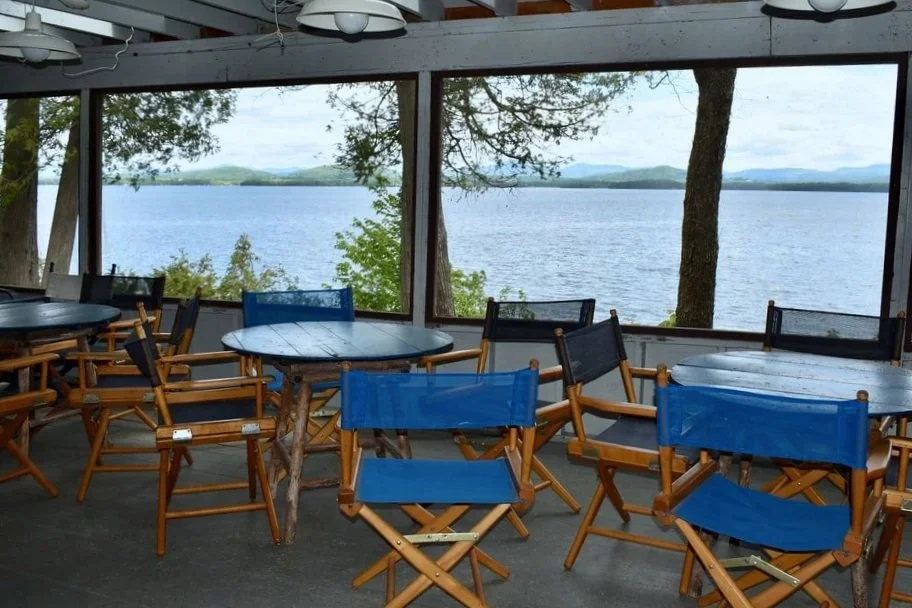 Indoor dining area with wooden tables and chairs, large windows with lake and trees outside, and mountains in the distance.