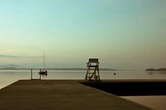 Lifeguard chair on a dock overlooking a calm body of water with a boat in the distance at sunset.