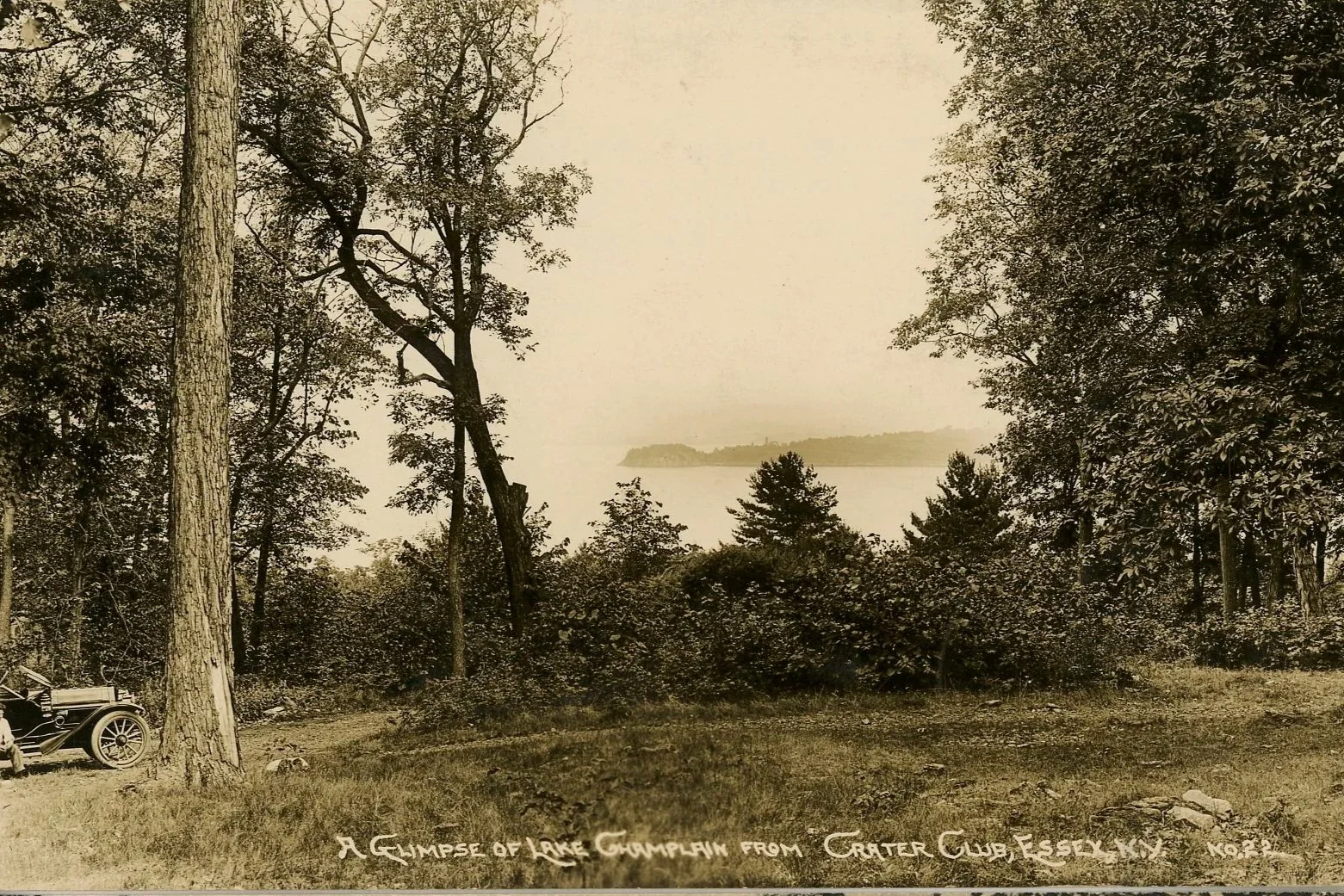 A sepia-toned photograph of a lakeside view through a wooded area with trees in the foreground, a body of water stretching to the horizon, and a distant island. A vintage car is parked on the left side. Handwritten text at the bottom reads "A glimpse of Lake Champlain from Crater Club, Essex NY."