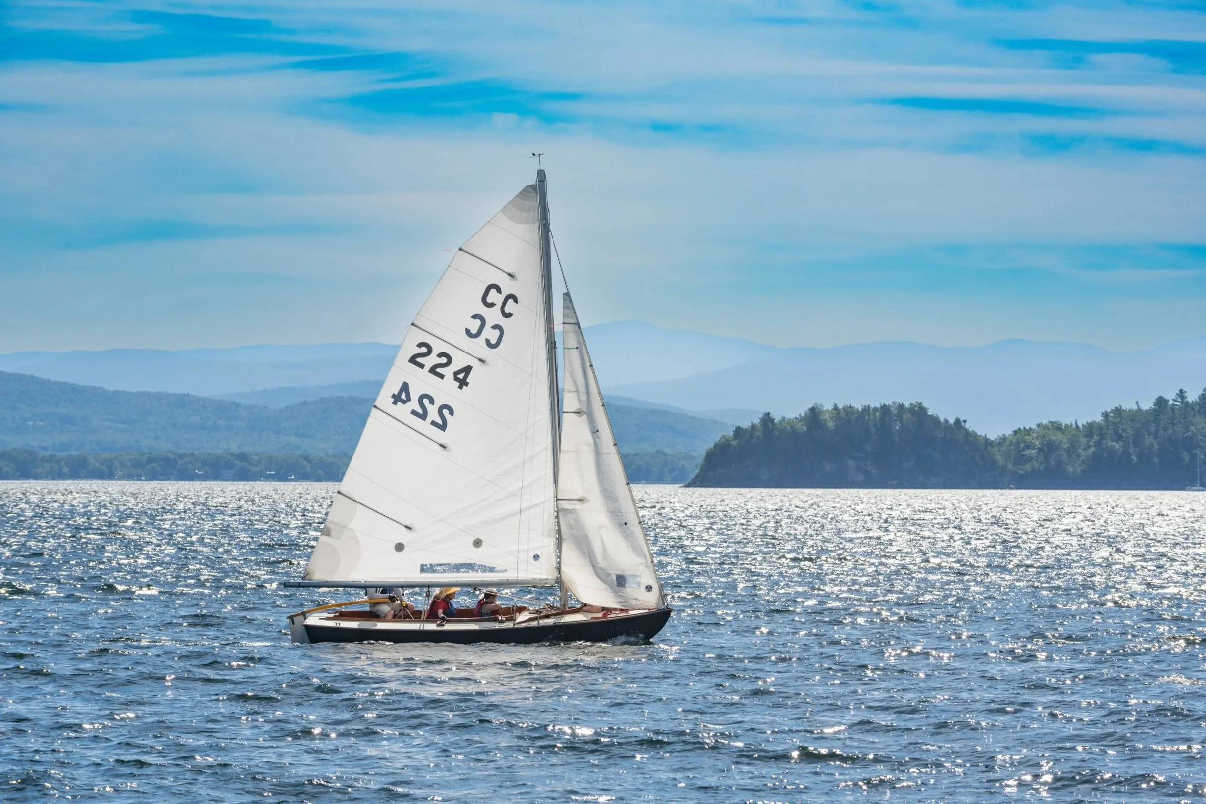 A sailboat with a white sail on a body of water, with a few people on board, and hills and mountains in the background under a partly cloudy sky.