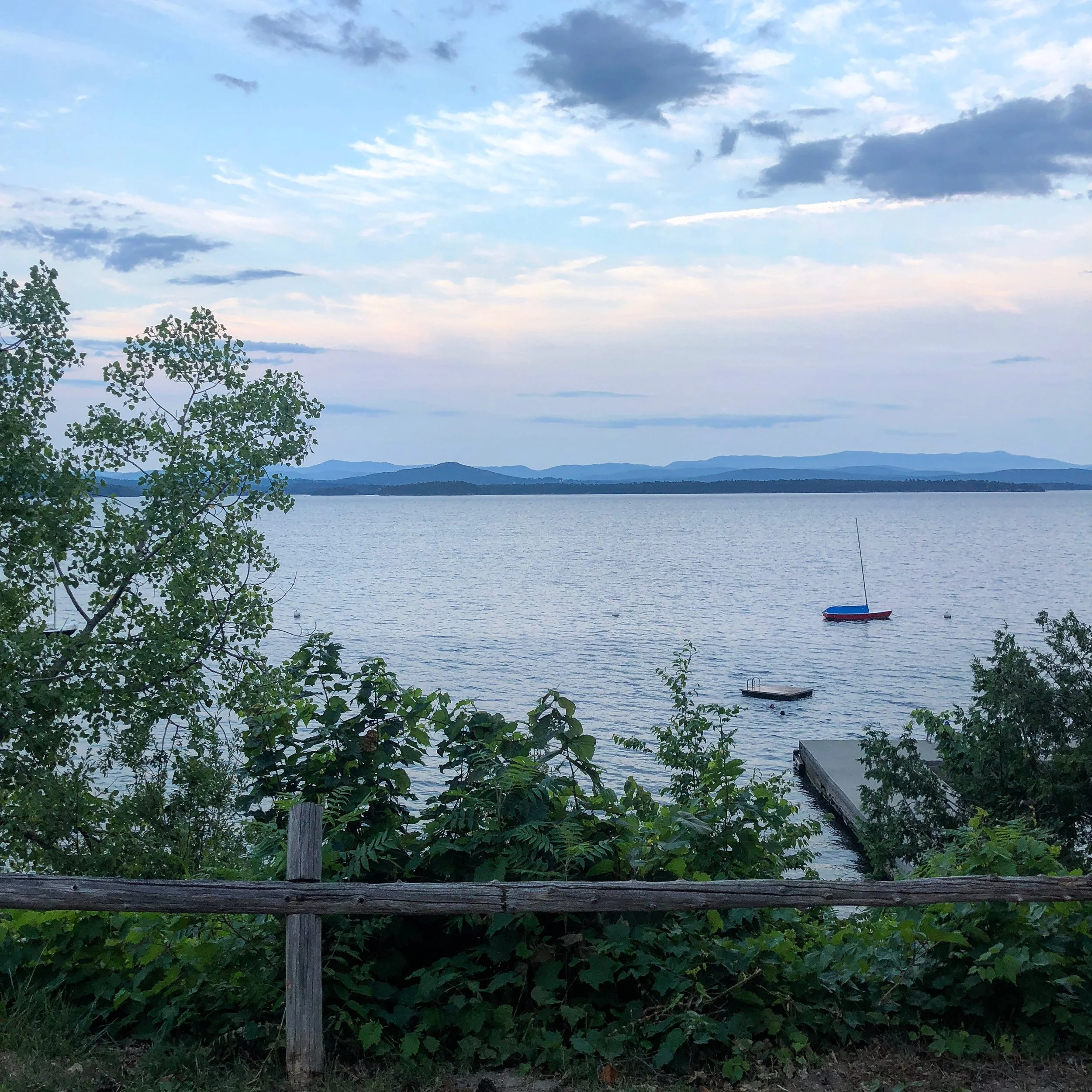 A scenic view of a large body of water with mountains in the distance under a partly cloudy sky, seen from a wooded area with leaves and a wooden fence in the foreground, and a small boat floating on the water.