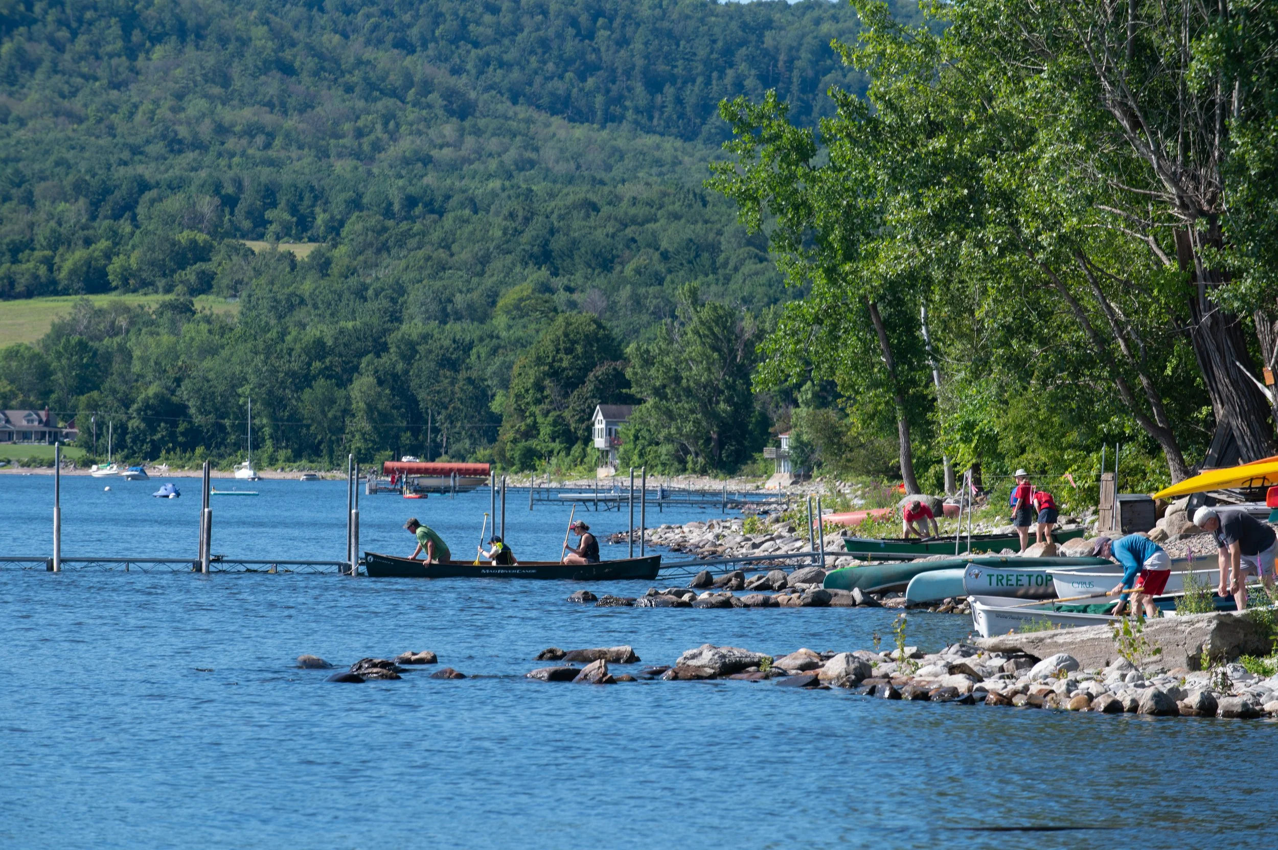 People near a lakeshore preparing small boats or kayaks, with trees and hills in the background.