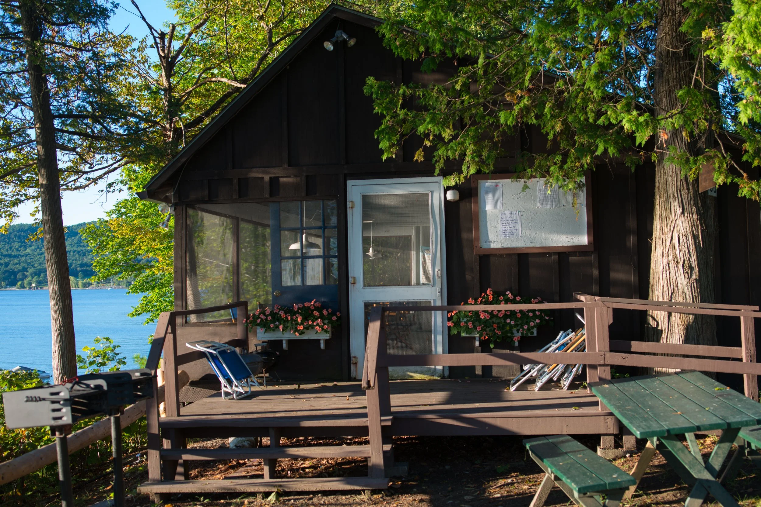 A small wooden lakeside cabin with black exterior walls and a glass door. The deck is decorated with potted pink flowers and has outdoor furniture, including a bench and a chair. Tall trees with green leaves partially obscure the cabin. The backgroun