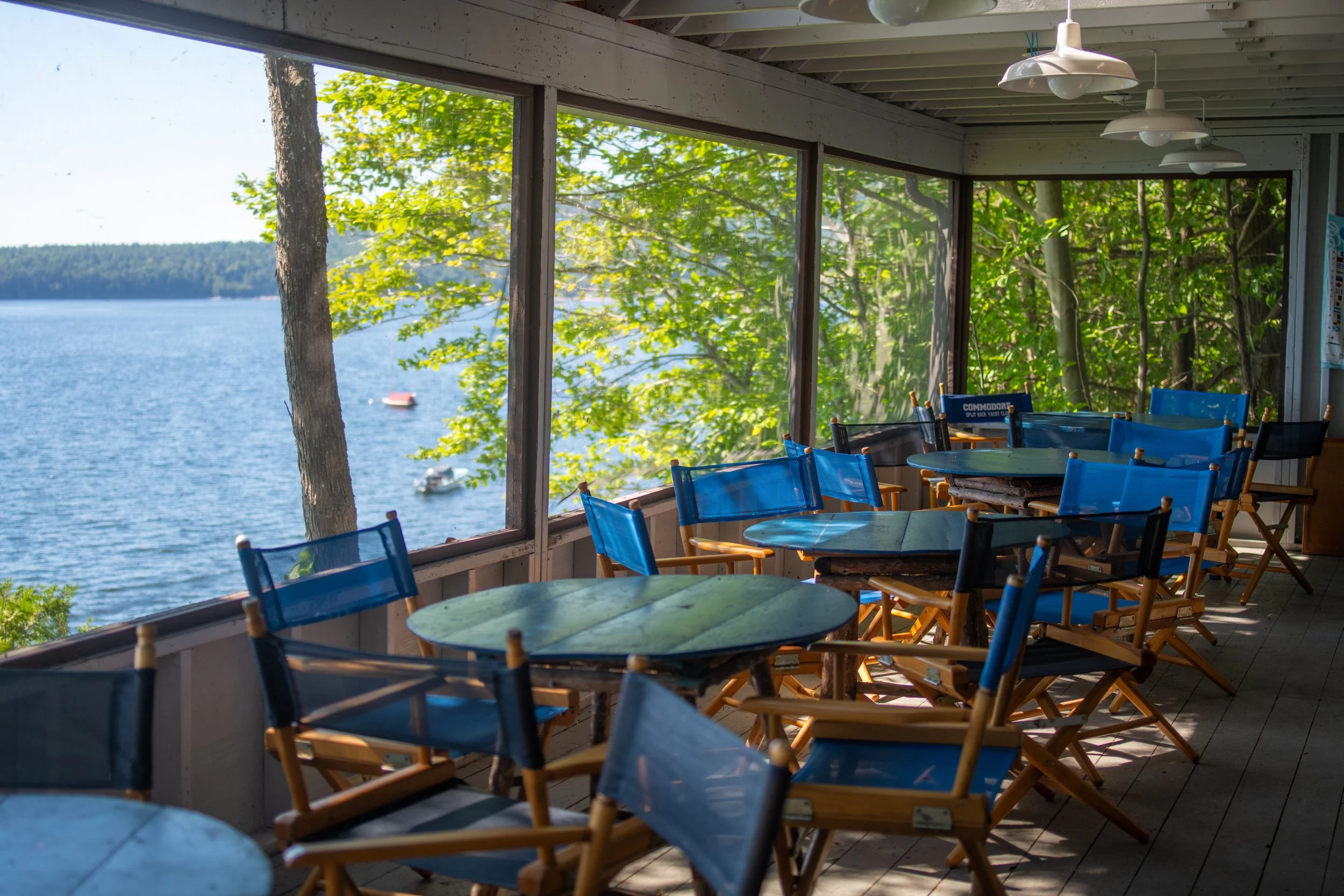 View of a lakeside scene through large windows, with blue chairs around tables inside a rustic room with exposed beams, overlooking a lake with a boat and trees on the shoreline.