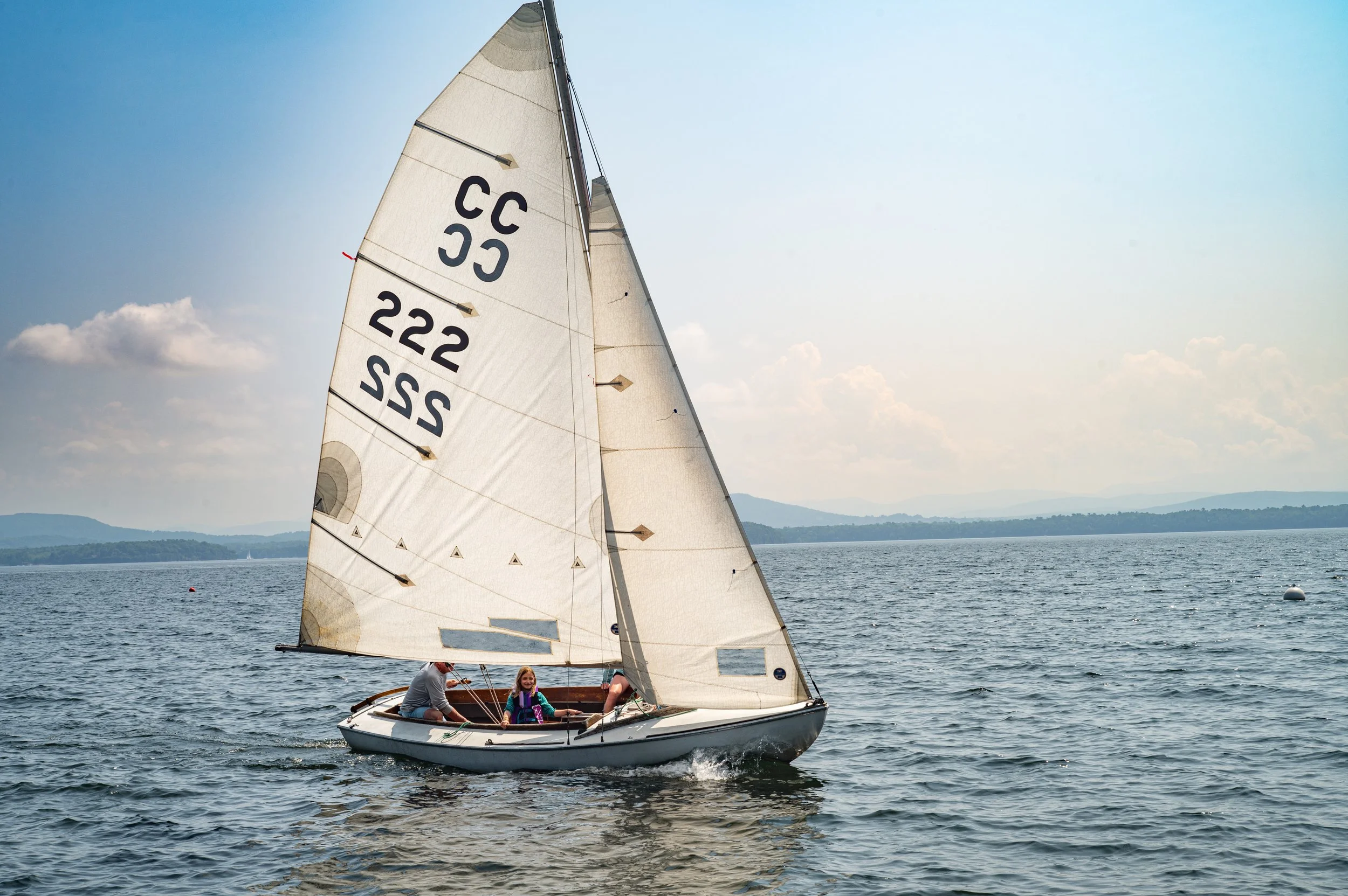 A sailboat with a white sail numbered 222 and 55, sailing on the water with a landscape of hills and a cloudy sky in the background. Two children are sitting in the boat, enjoying the trip.