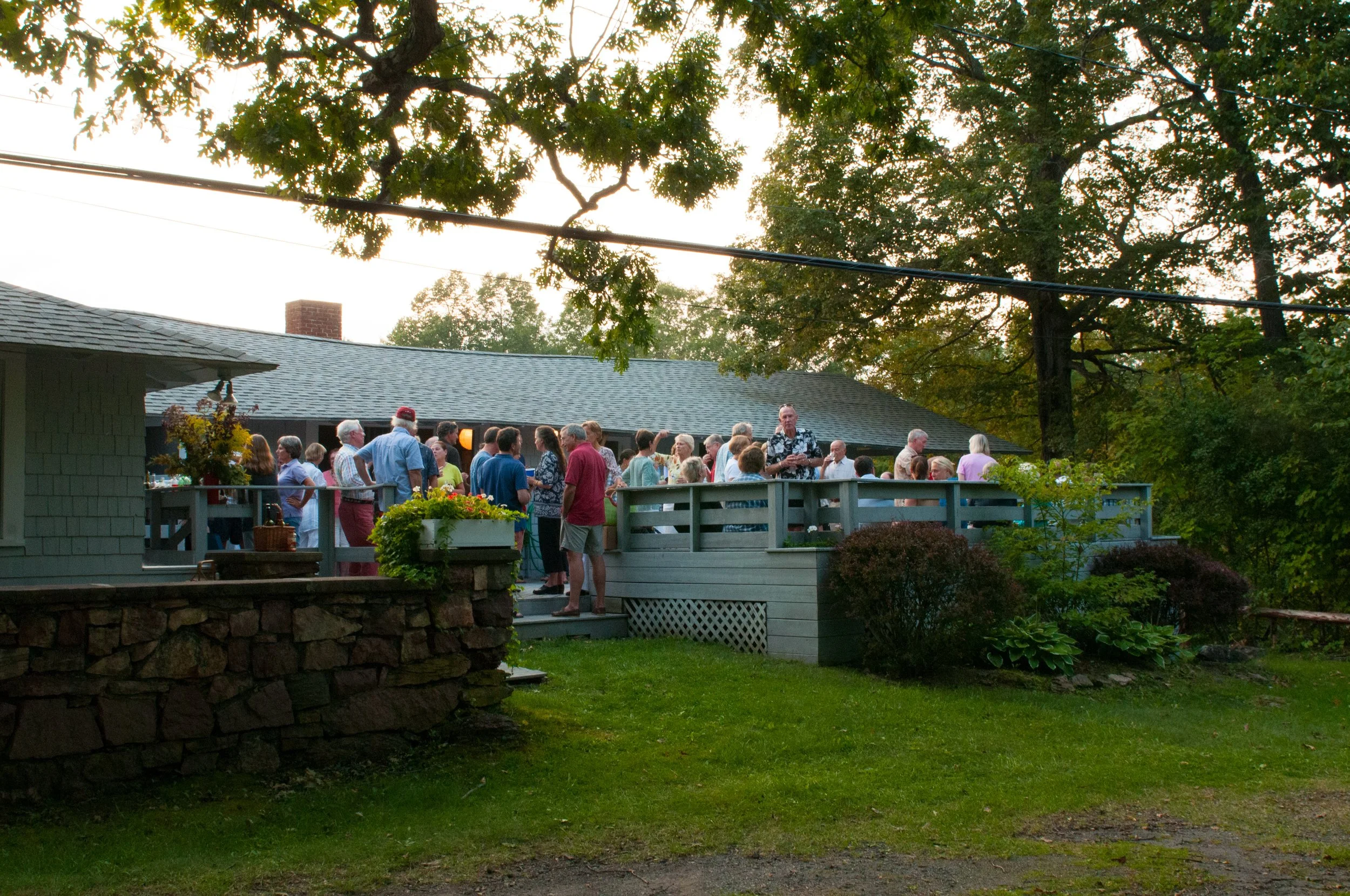 A backyard gathering with people socializing on a deck, surrounded by trees and greenery at sunset.