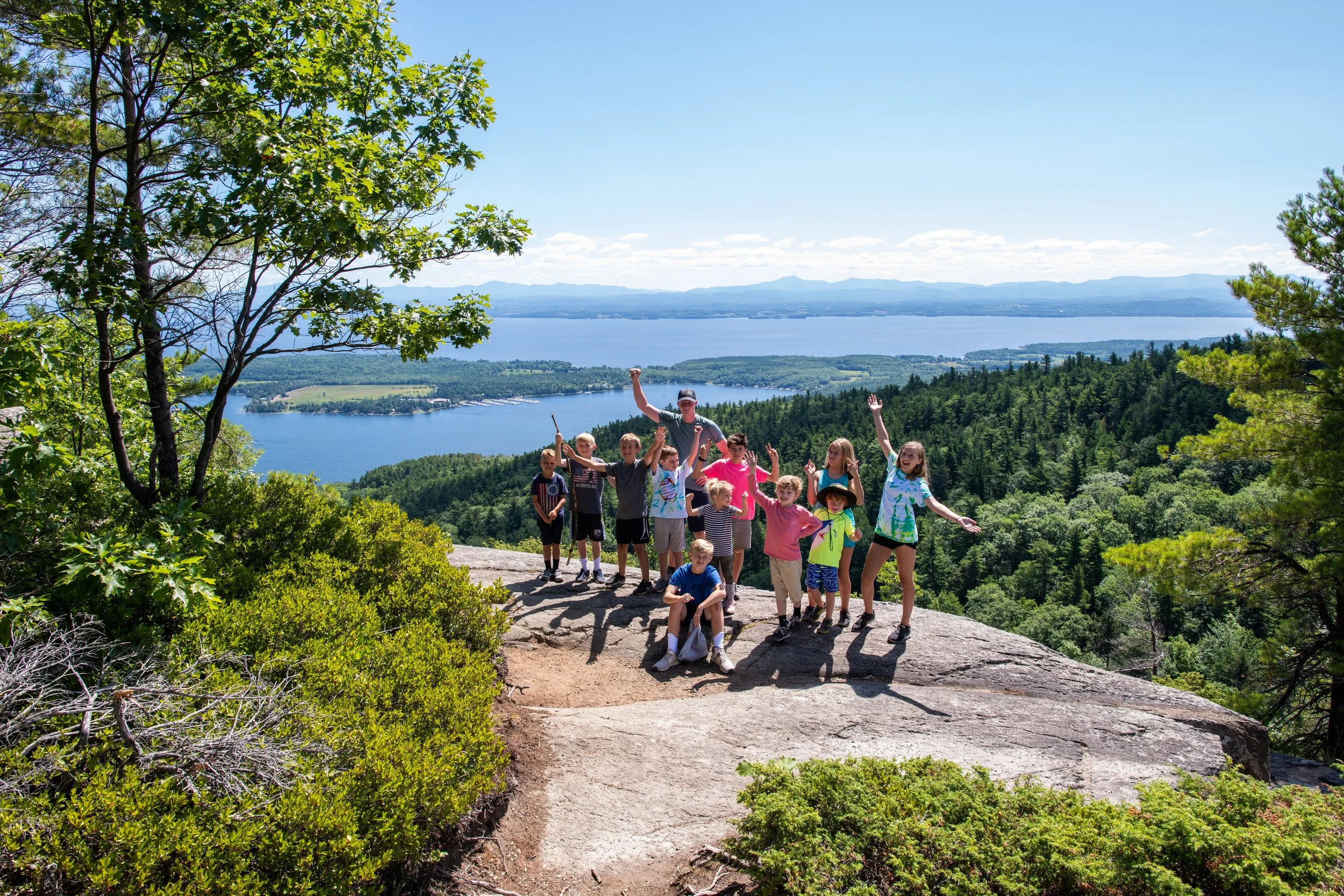 A group of children and an adult standing on a rocky outcrop on a hiking trail, overlooking a large lake with forested shores and mountains in the distance, on a sunny day with a clear blue sky.