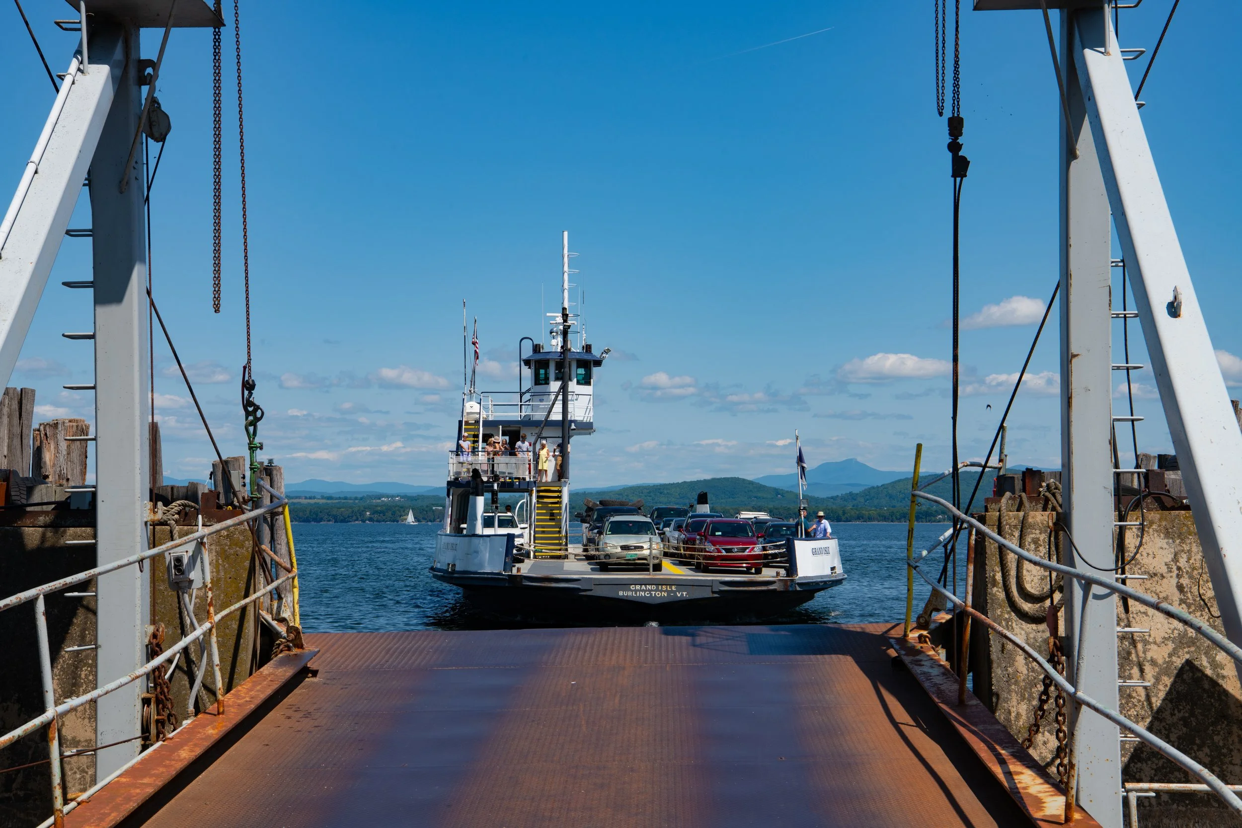 View of a ferry dock with cars on the vehicle deck, seen through metal railings and chains. In the background, a ferry boat is approaching or departing from a harbor on a clear, sunny day with a blue sky and distant mountains.