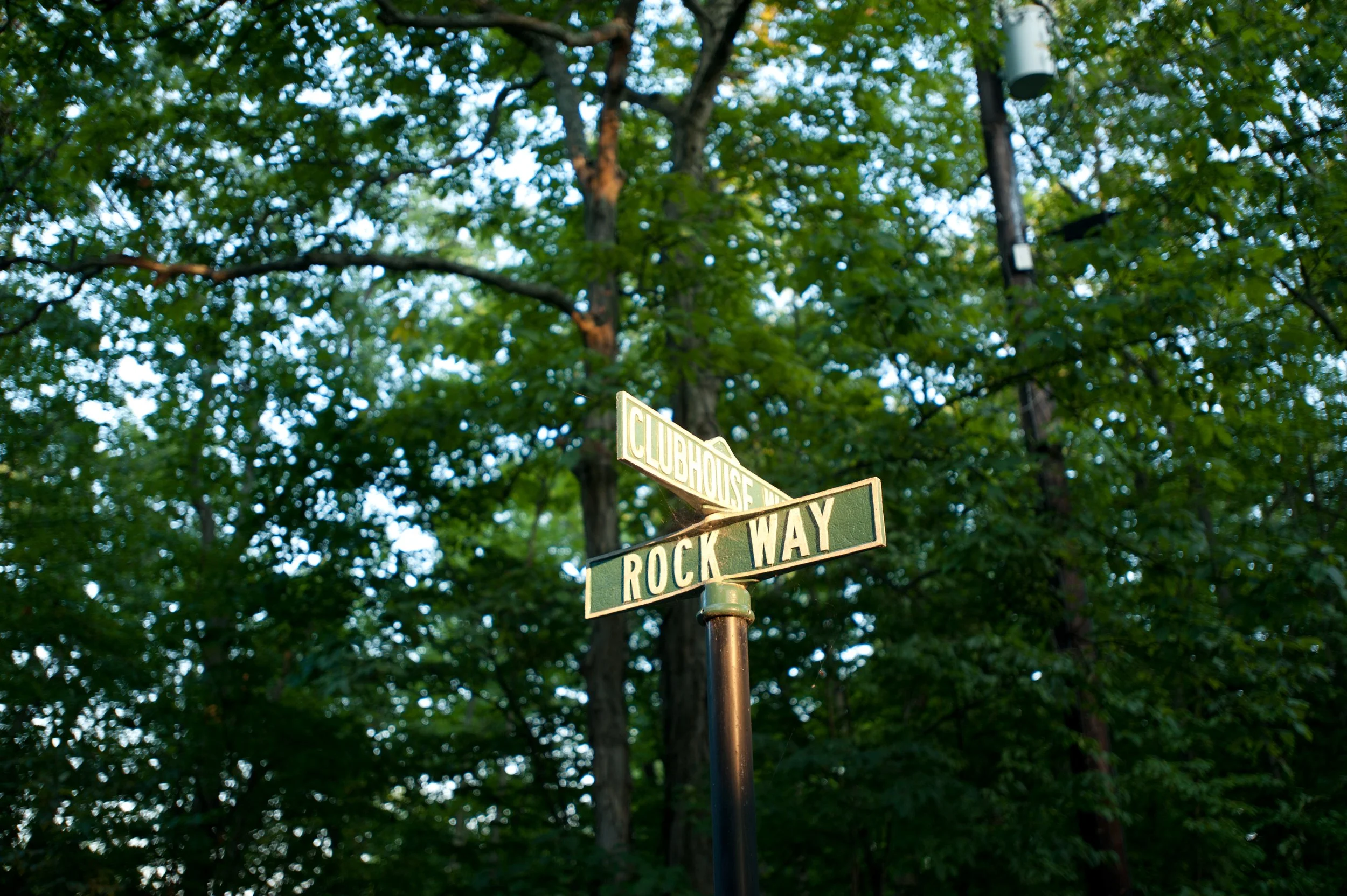 Street sign at a corner with the names 'Clubhouse Way' and 'Rock Way' against a backdrop of green trees and a clear sky.