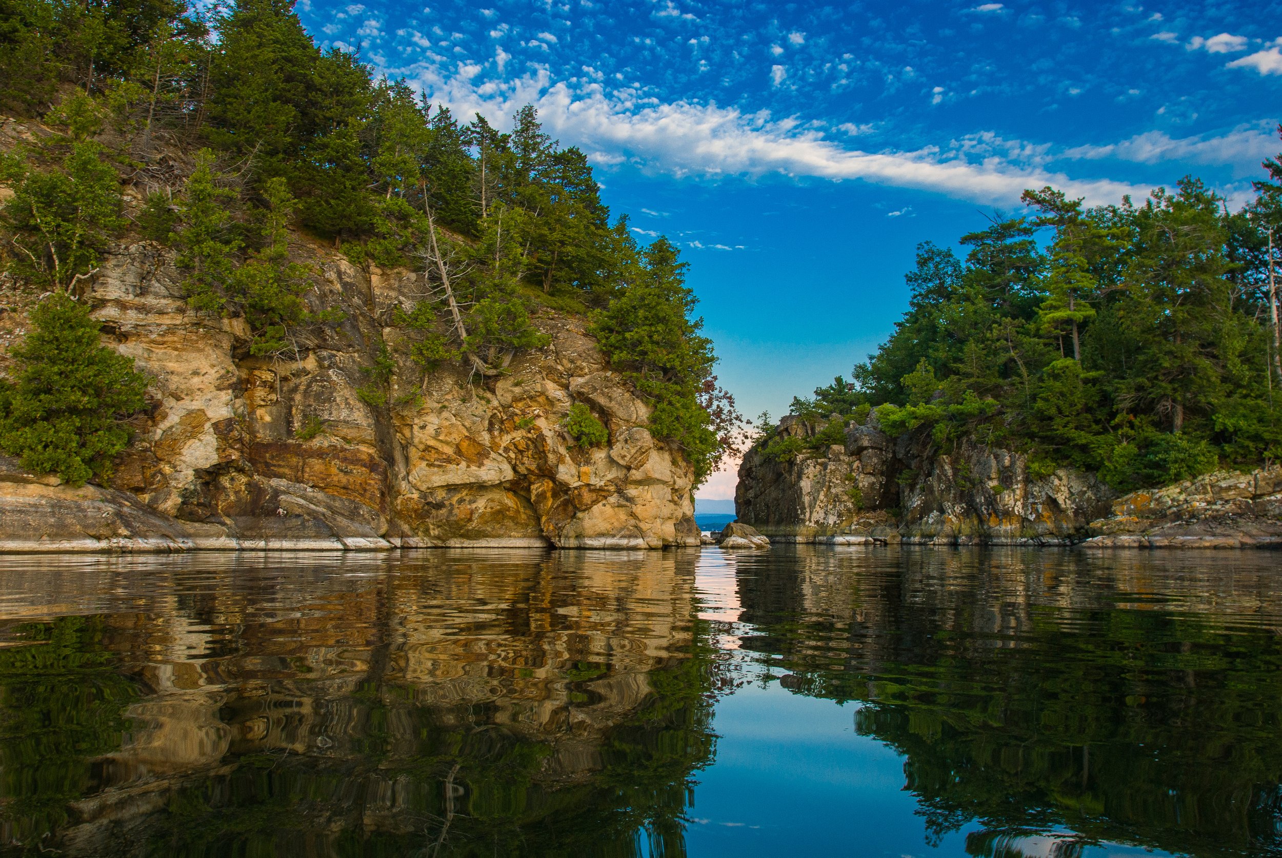 A serene river flows between rocky cliffs covered with green trees under a partly cloudy sky.