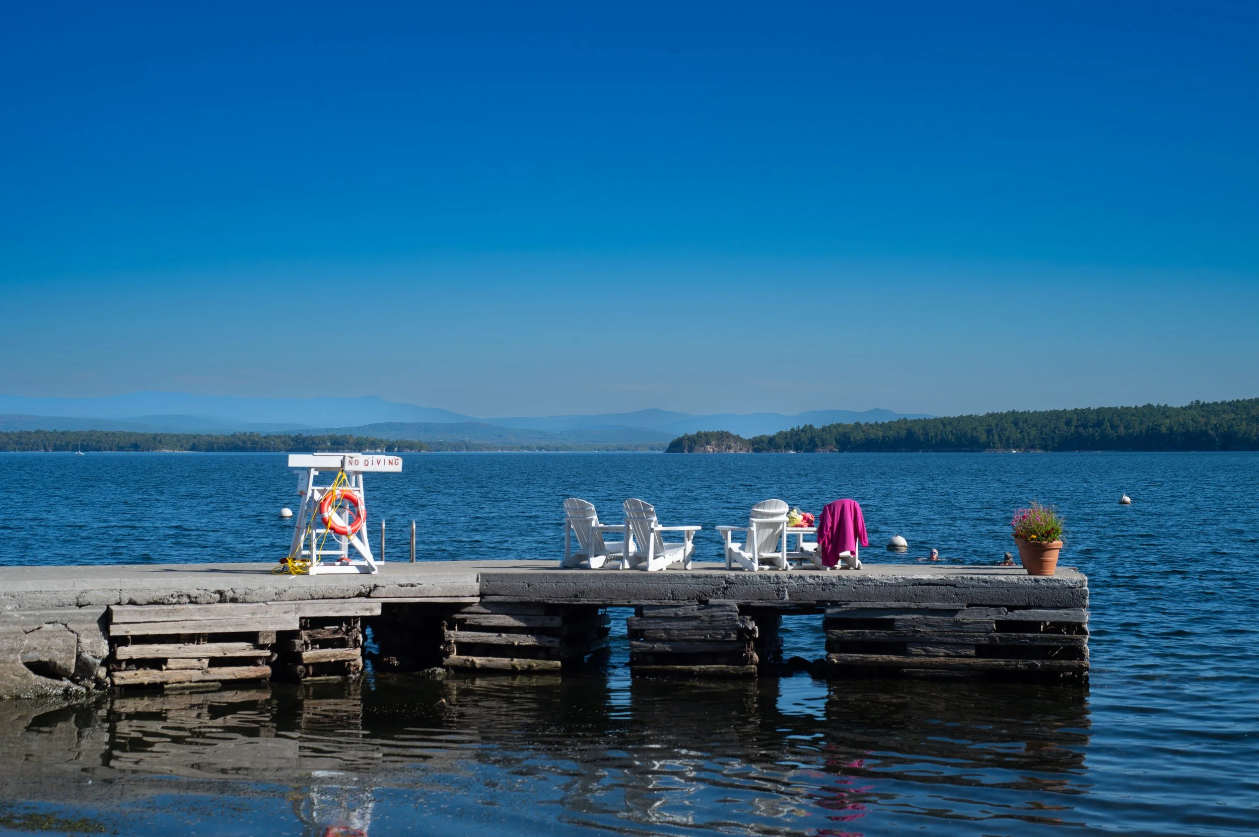 A lakeside scene with a wooden dock extending into calm water, featuring white lounge chairs and colorful towels. In the background, there are distant mountains under a clear blue sky.