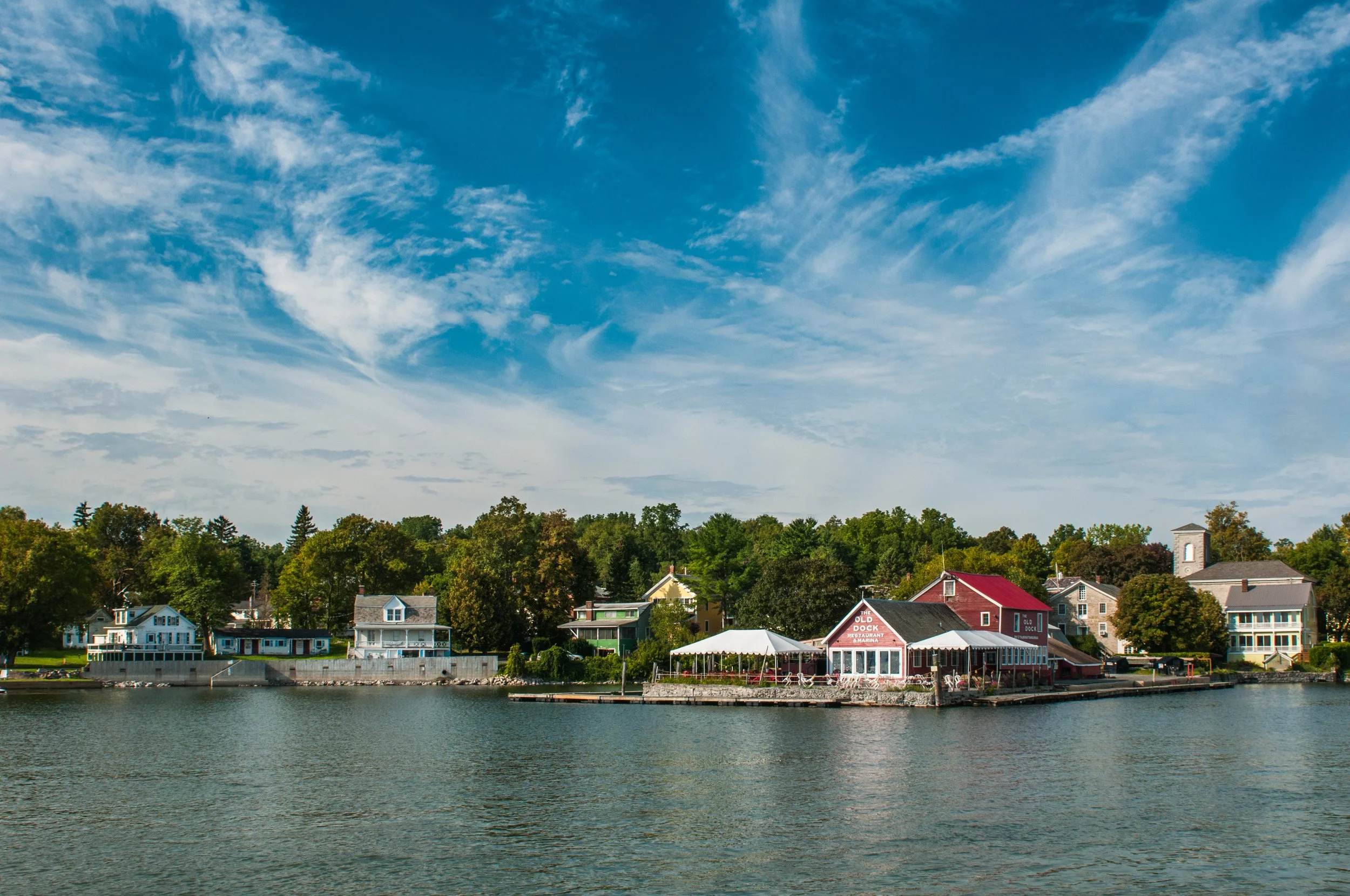A waterfront scene with houses and buildings along the shoreline, a railing, and trees in the background under a partly cloudy sky.