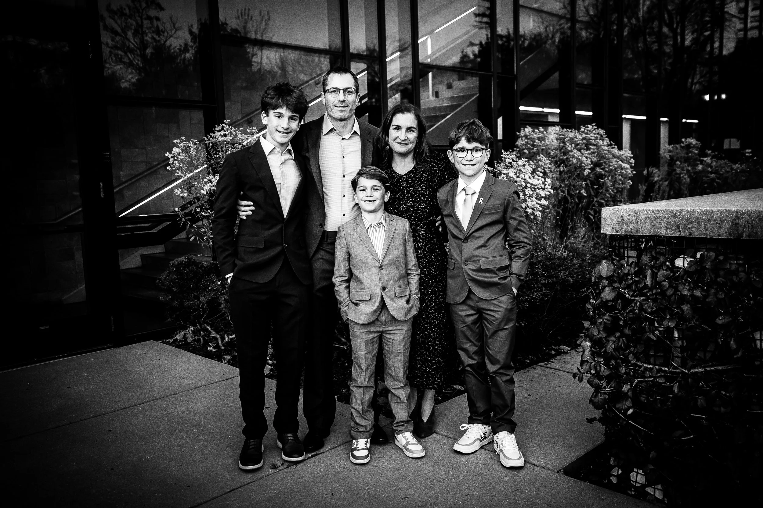 A family of six, including three boys and a woman, pose for a group photo outside a modern building with glass walls, at dusk. All are dressed in formal attire, with some wearing suits and ties.