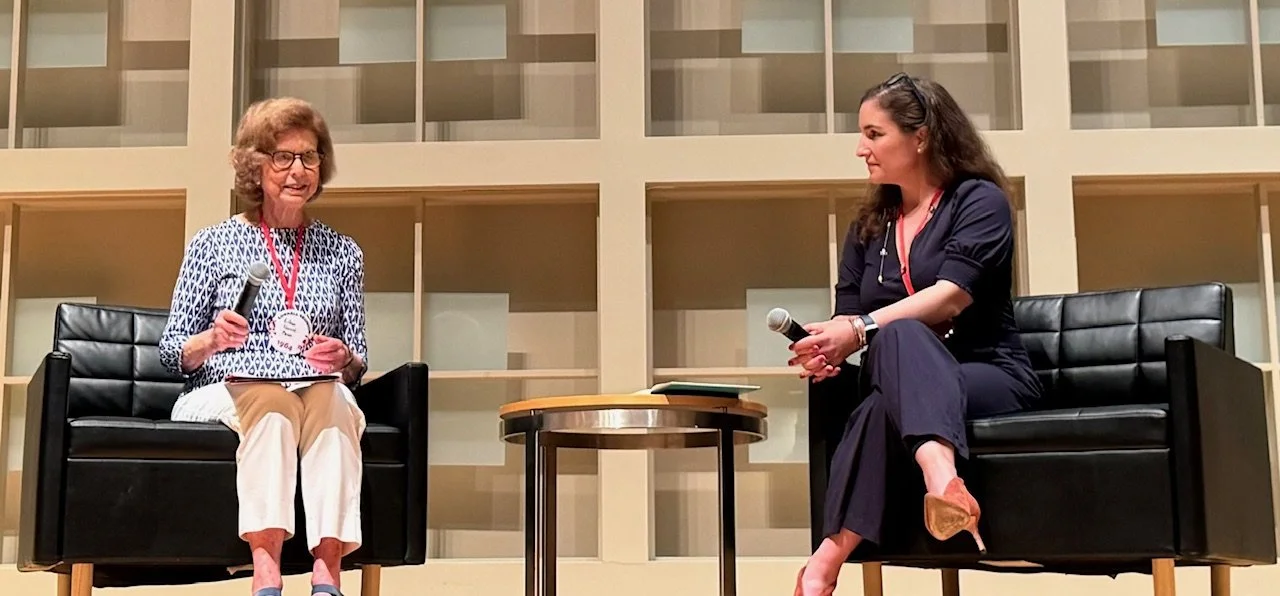Two women sitting on black armchairs on a stage, holding microphones, engaging in a conversation, with a modern beige background and a small round table between them.