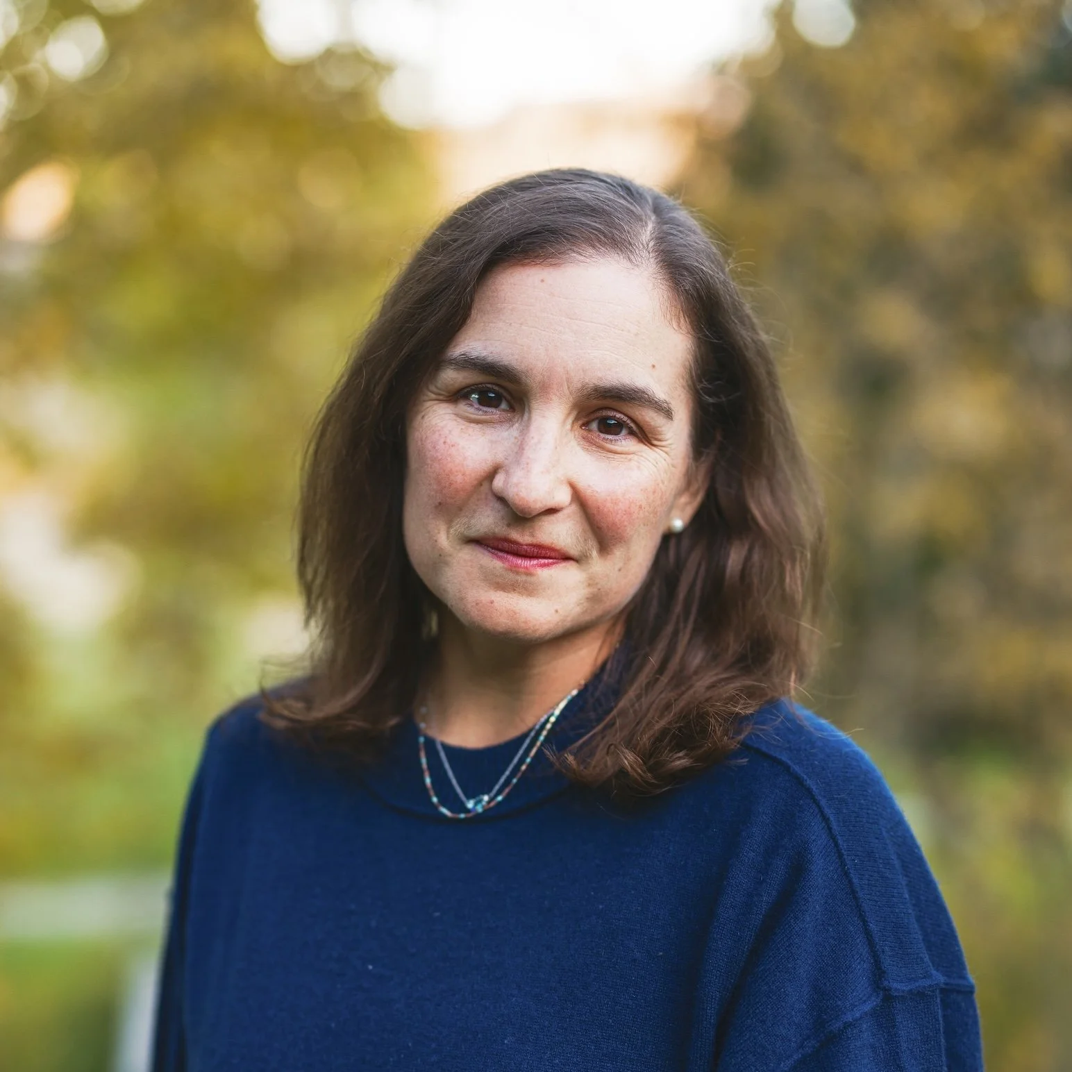 Katie Dealy headshot. A woman with shoulder-length brown hair, wearing a navy blue sweater and layered necklaces, standing outdoors with a blurred background of trees and foliage during fall.
