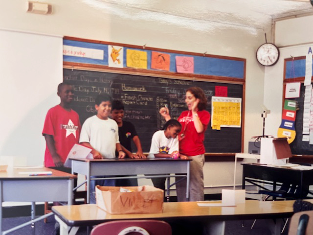 Katie Dealy in her classroom. A teacher and four students in a classroom standing at a desk, with a blackboard behind them and colorful posters above.