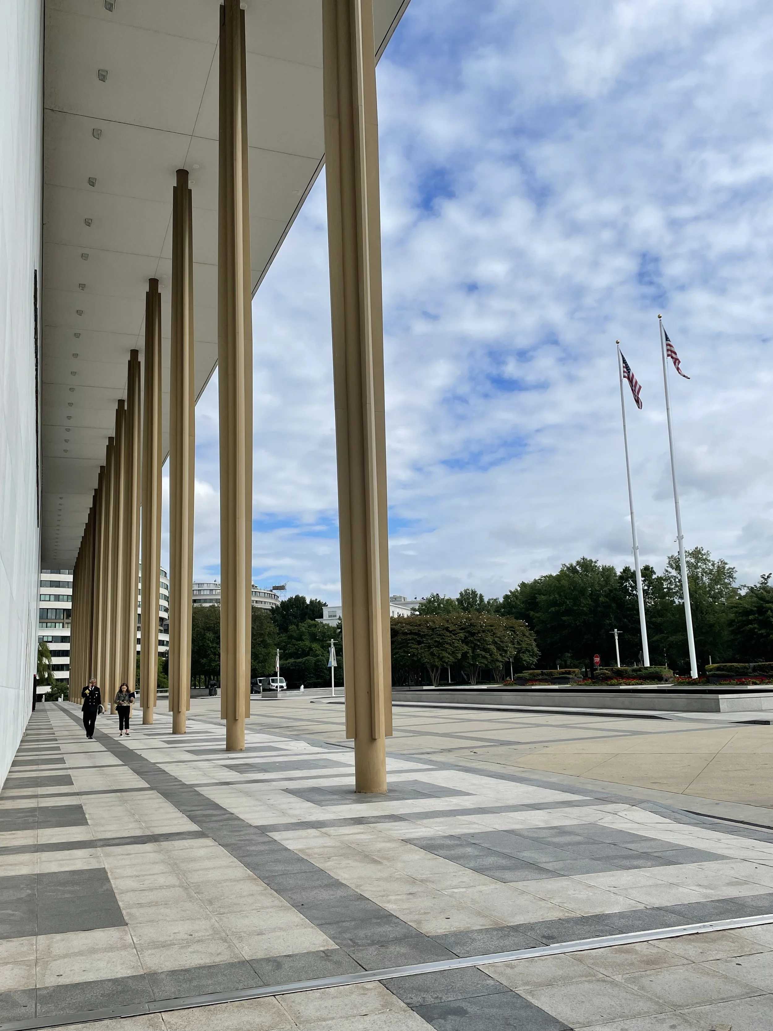 Katie Dealy walking with U.S. Surgeon General Vivek Murthy outside the Kennedy Center. Empty outdoor plaza with tall beige pillars supporting a modern building, two flagpoles with American flags, and trees in the background under partly cloudy sky.