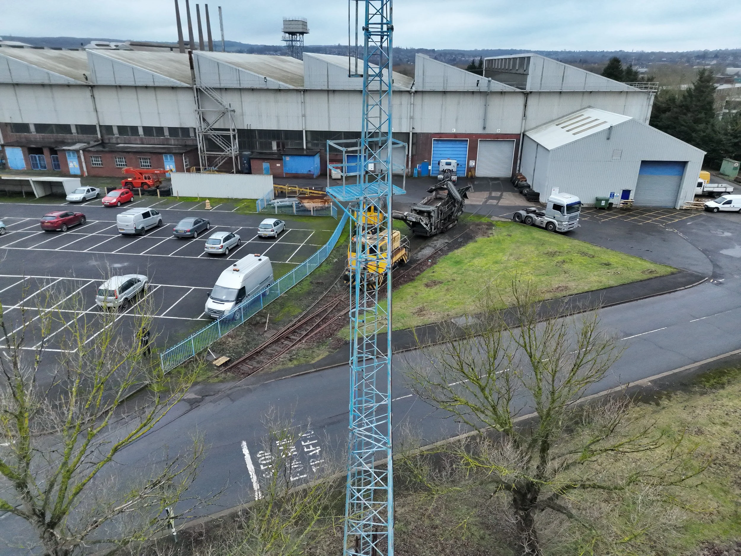 An industrial building with a parking lot and a railway track, construction equipment, and a partly cloudy sky.
