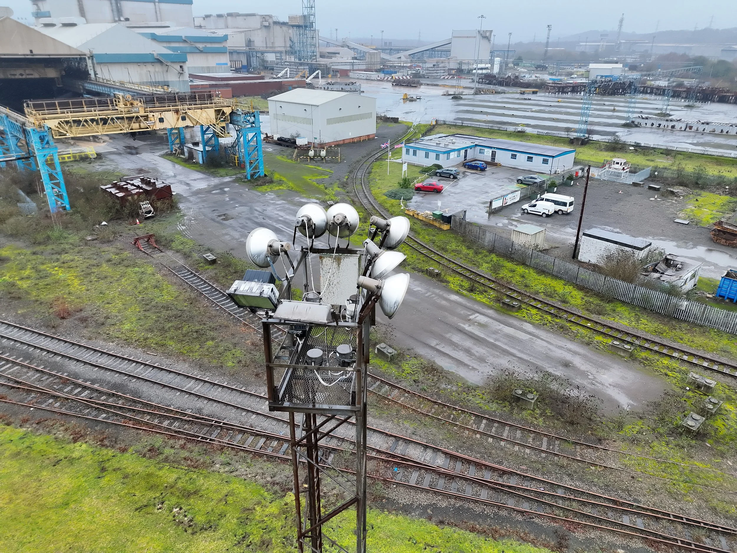 Overhead view of an industrial port area with train tracks, a parking lot with cars, warehouses, and machinery, under a cloudy sky.