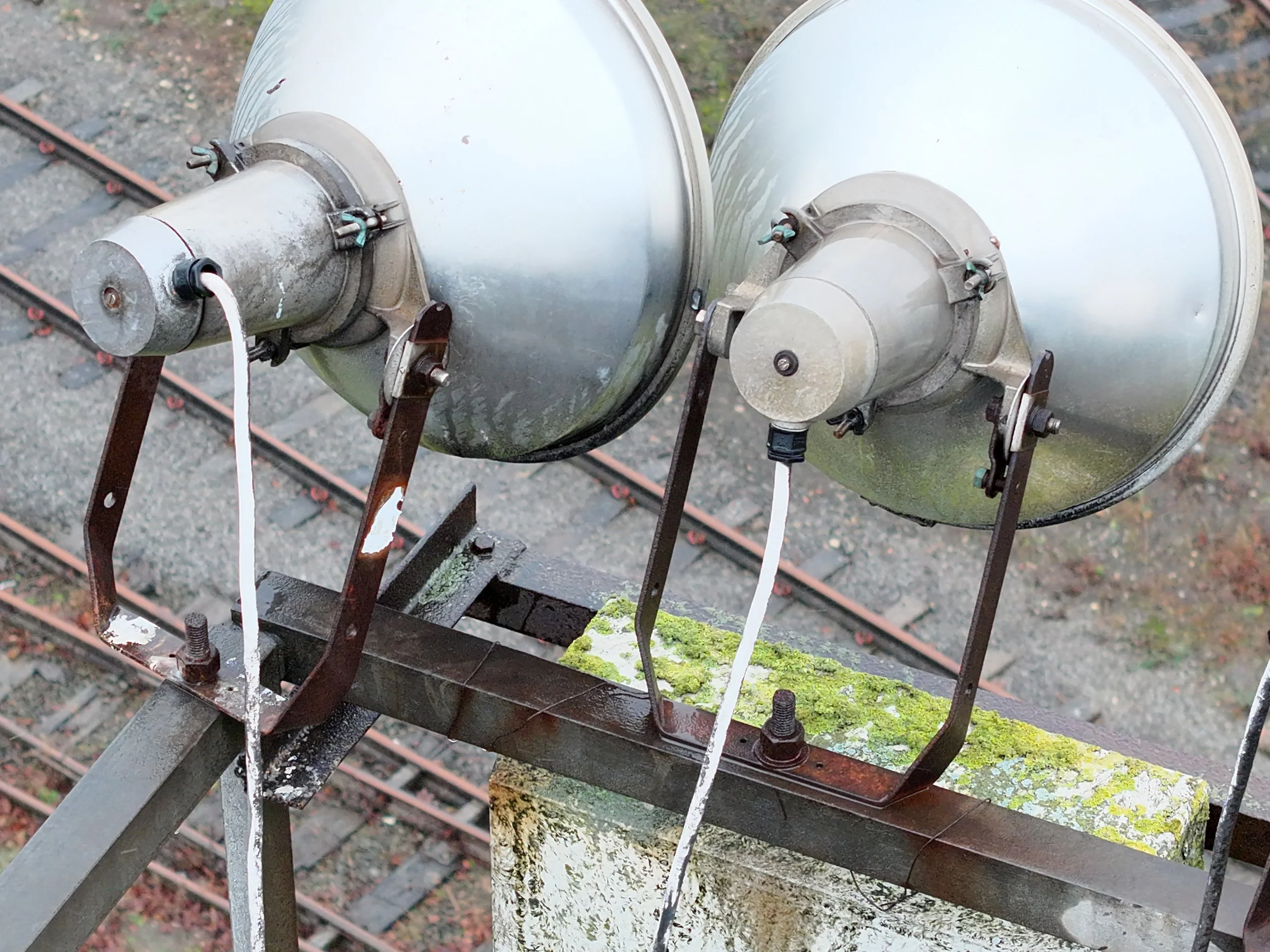 Two large metallic antennas or satellite dishes mounted on a rusted metal structure above a moss-covered concrete block, with train tracks visible below.
