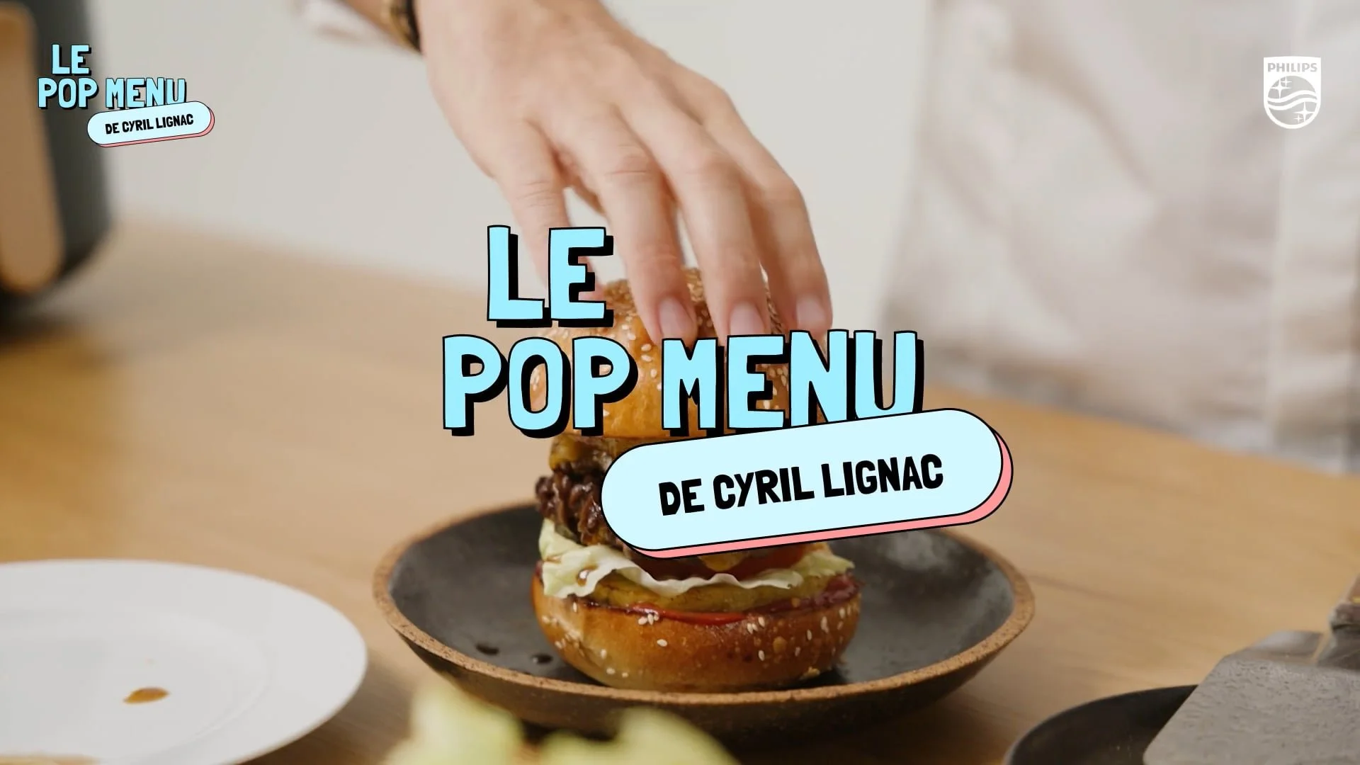 Un sandwich avec de la viande, de la salade, dans un bun et une main qui le tient, sur une assiette noire. Textes en français indiquant "Le pop menu de Cyril Lignac".