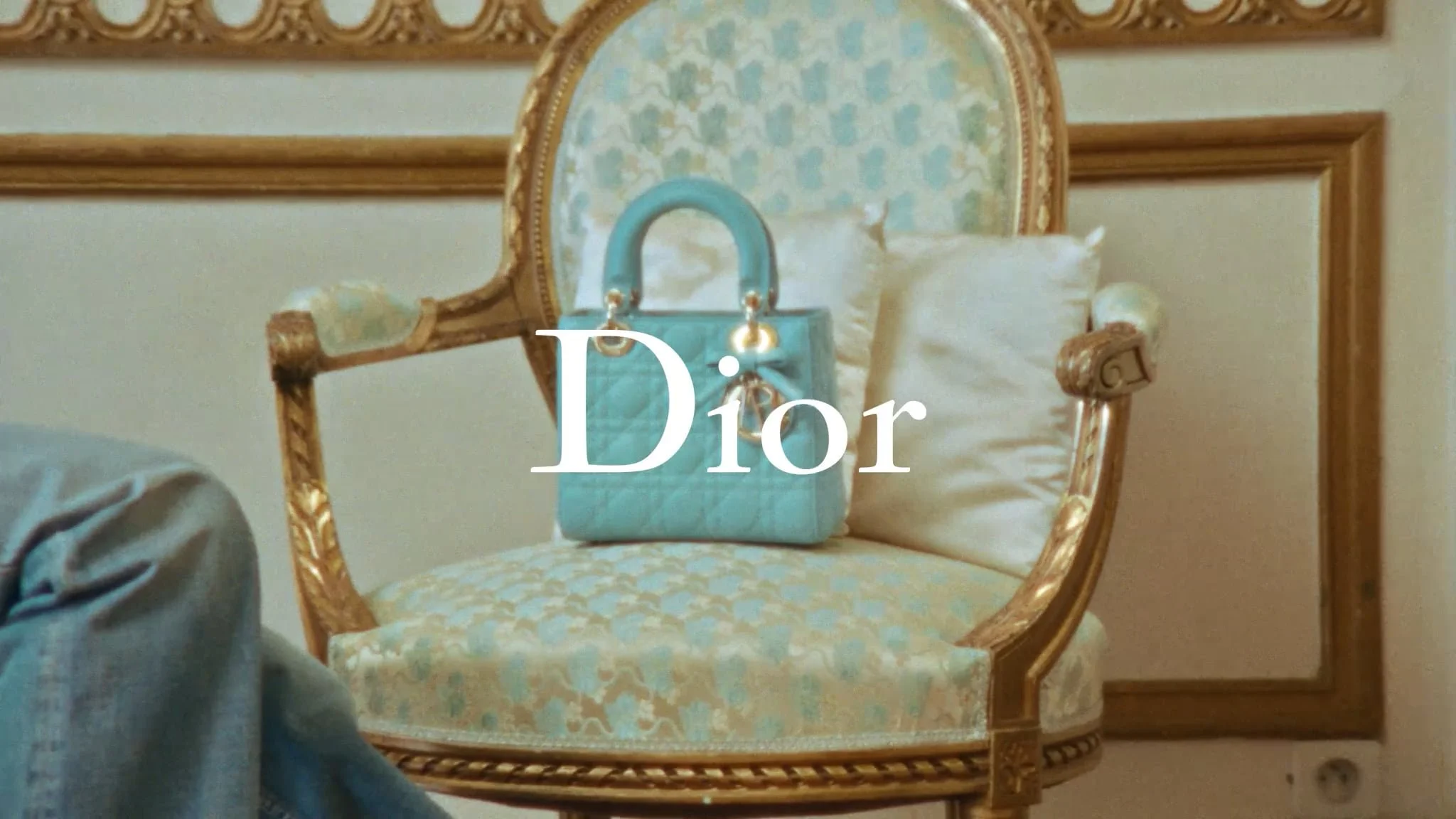 Un fauteuil ancien avec un tissu à motifs, un coussin blanc et un sac à main bleu posé dessus, avec le logo Dior en surimpression.