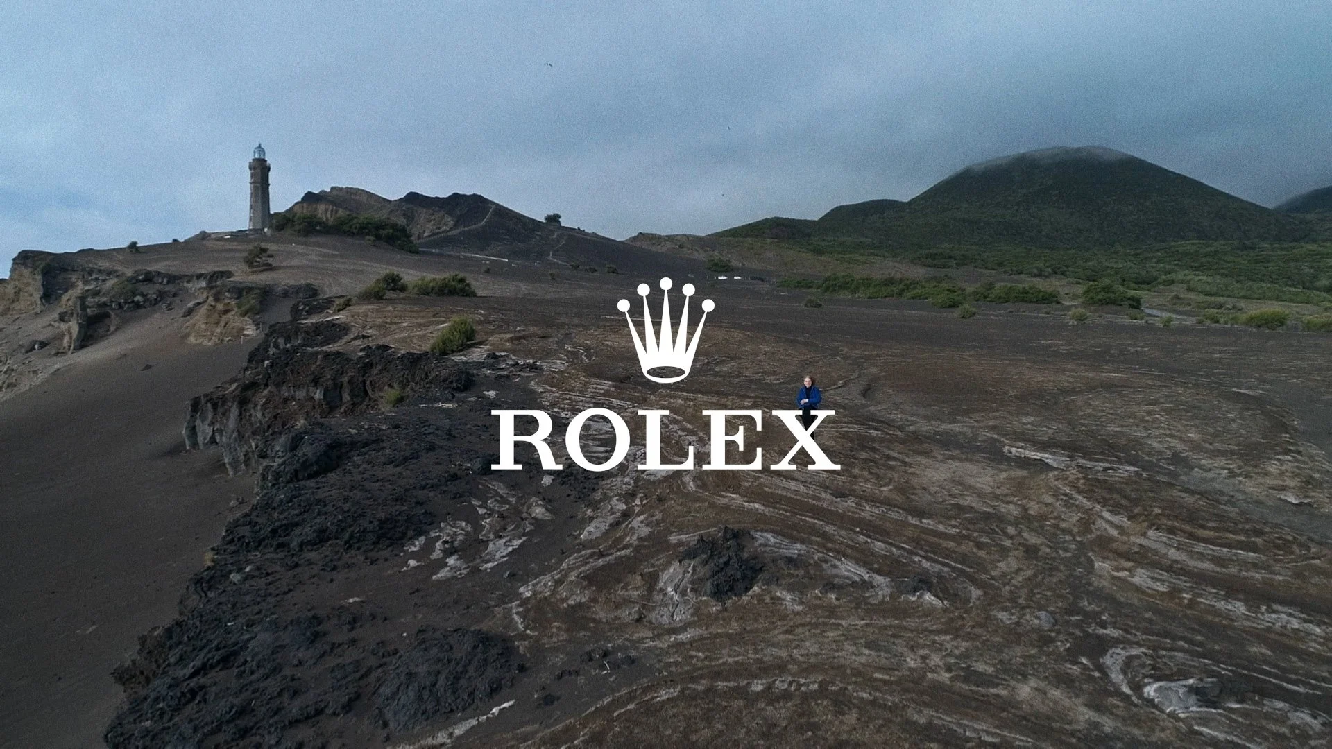 Paysage de volcans avec un phare et un sentier, logo Rolex superposé au centre.