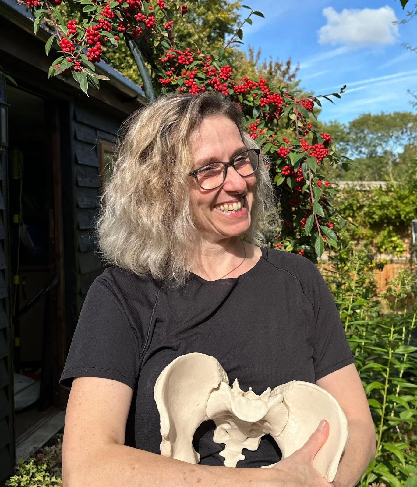 A woman with curly blonde hair and glasses smiling while holding a large anatomical skull model in front of a garden with red berries and green foliage under a partly cloudy sky.