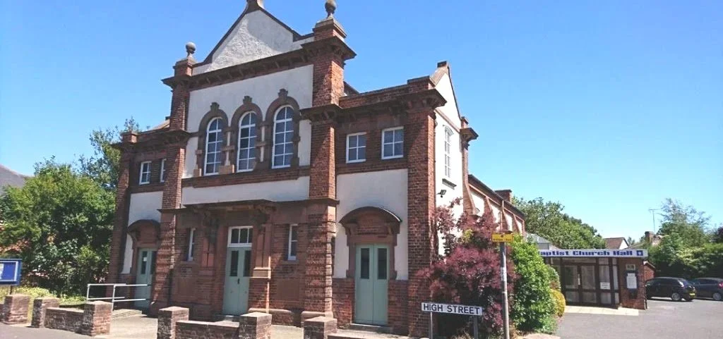 A multi-story brick church building with arched windows, a small shop or office with a sign that says 'Baptist Church Mail', trees, a street sign that says 'High Street', and a parking lot on a sunny day.