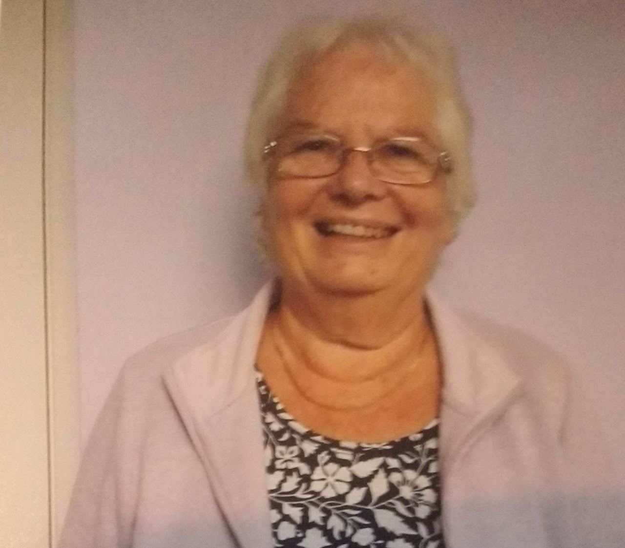 Smiling woman with glasses, short gray hair, wearing a patterned top and light-colored jacket, standing against a plain wall.