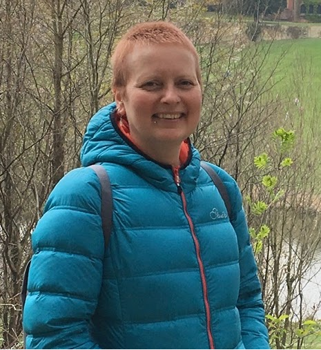 Person with short red hair wearing a blue puffy jacket outdoors, smiling, with bushes and trees in the background.