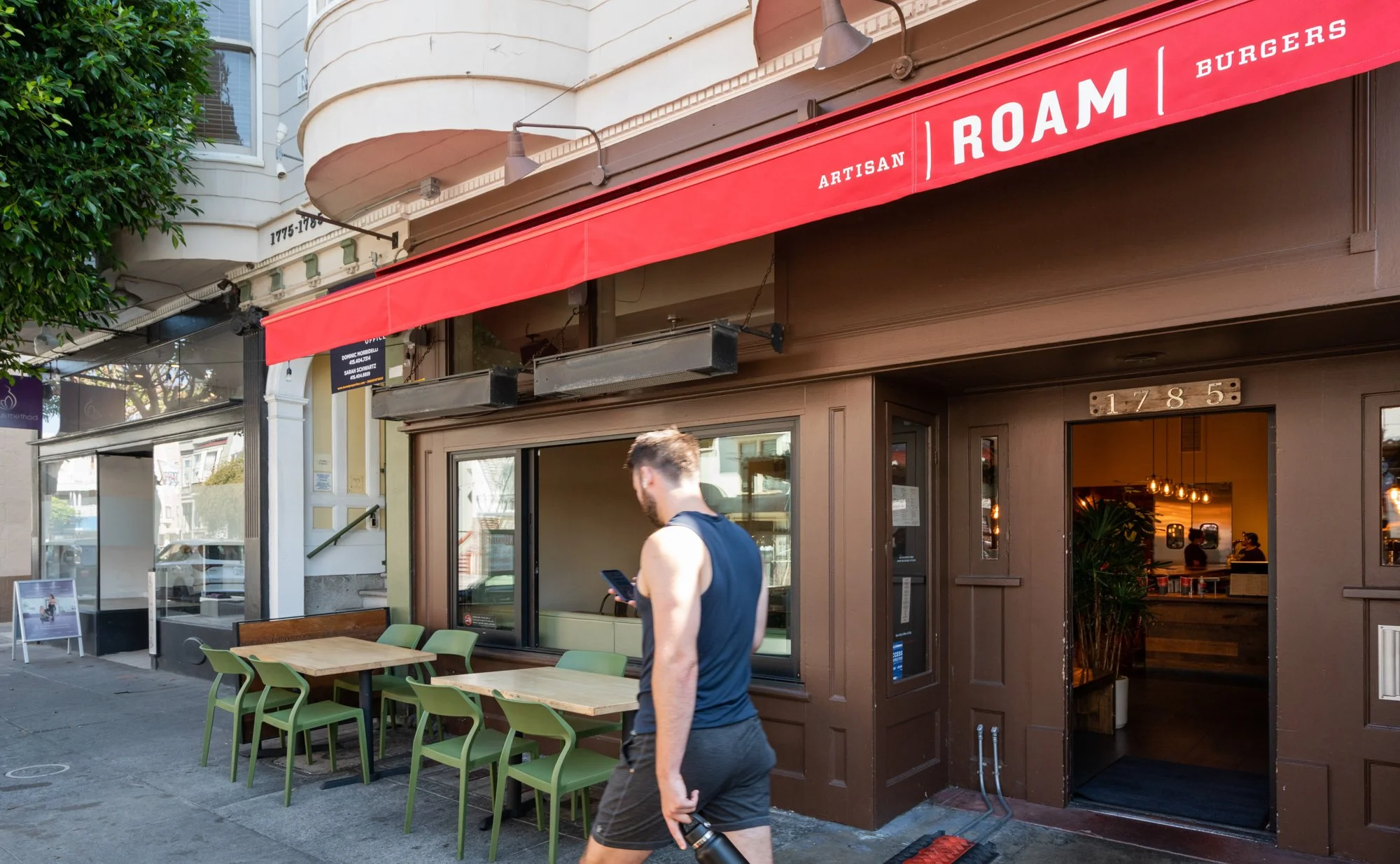 Man in sleeveless shirt and shorts walking past a restaurant with outdoor seating, red awning reading "ARTISAN ROAM BURGERS," brown exterior, open door revealing interior with plants and hanging lights.