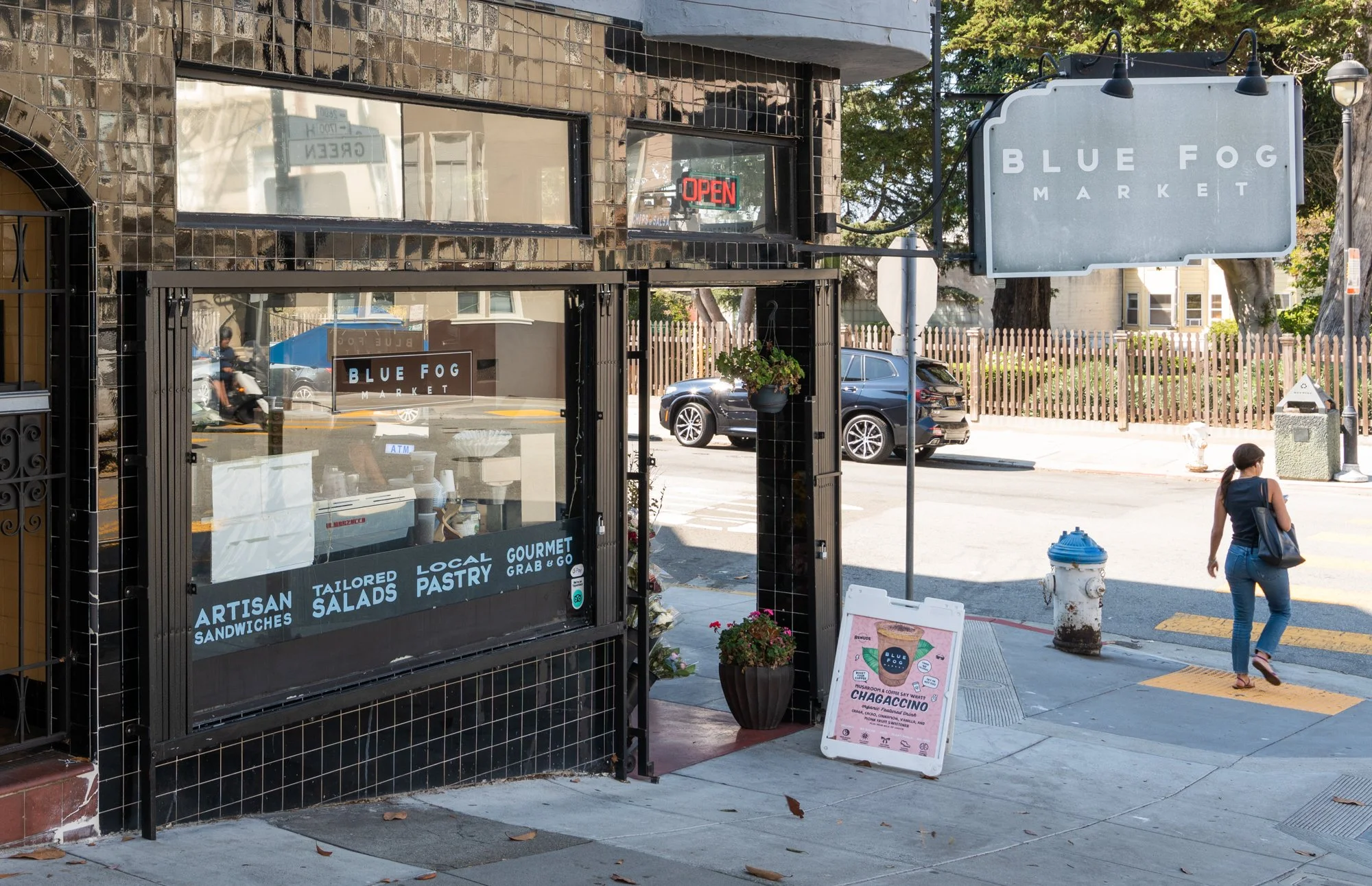 Street view of a small market called Blue Fog Market, with a black exterior and a window displaying signs for roasted sandwiches, salads, pastry, and grab-and-go options. A sidewalk chalkboard sign advertises chagaccino, and a woman with a bag walks 
