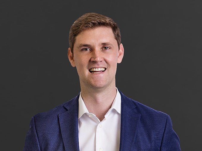 A young man with brown hair wearing a white shirt and a blue blazer, smiling against a gray background.