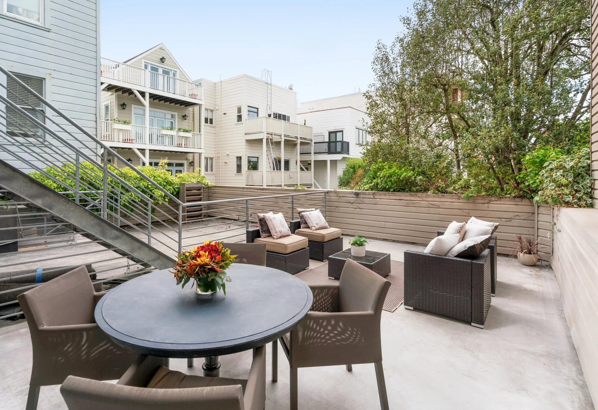 Outdoor patio with round table, chairs, and cushioned seating area enclosed by a wooden fence, surrounded by neighboring buildings and trees.
