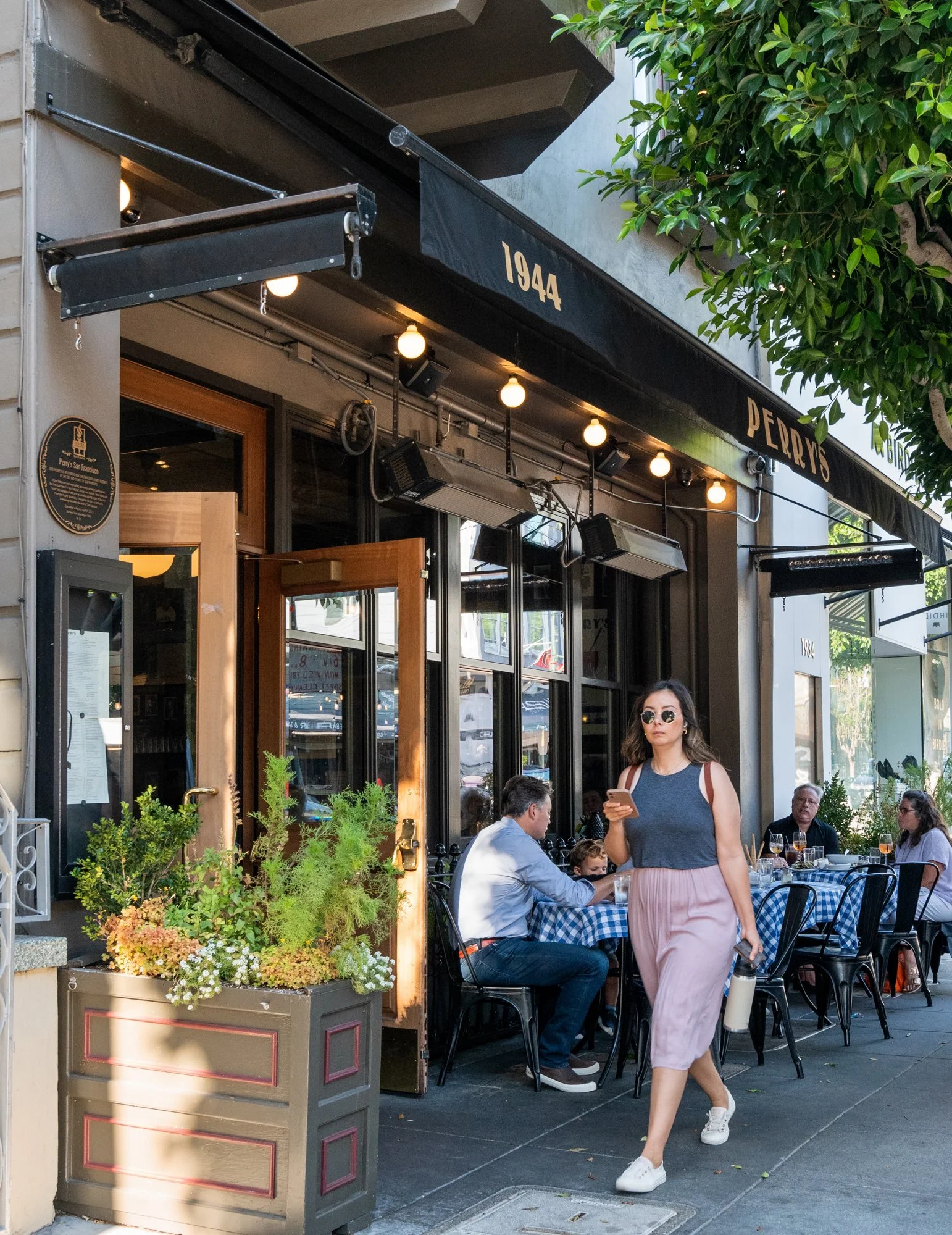 People dining outside Perry's restaurant with black awning named 1944, and a woman walking past holding a phone and a water bottle.