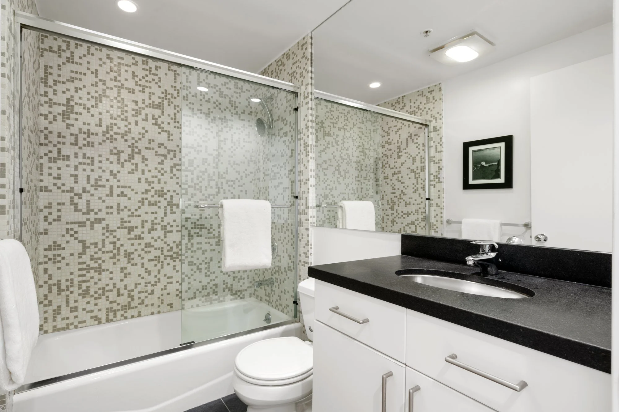 Modern bathroom with a glass-enclosed shower, white toilet, black countertop sink, white vanity cabinet, white towels, and a black picture frame on the wall.